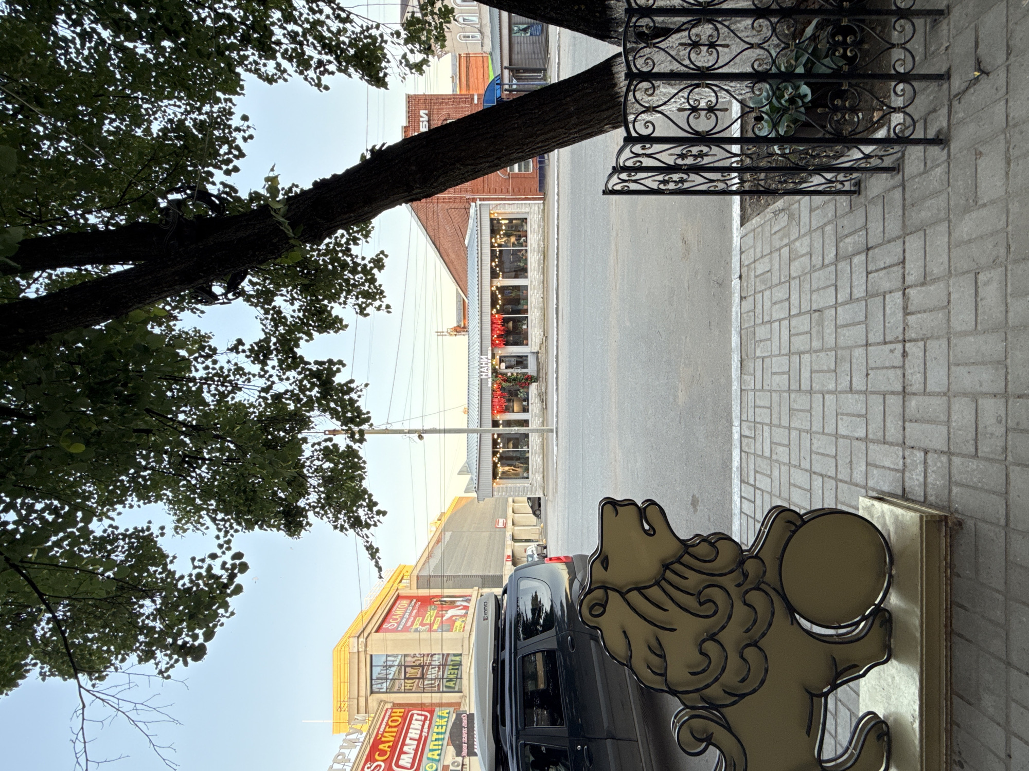 The image shows a street scene in Votkinsk, Russia. In the foreground, a golden lion statue with a ball stands on a paved walkway. To the left, a dark SUV with a roof carrier is parked. Across the street, several buildings are visible. On the left, a yellow building with signage for "САЙТОН" (SAITON) and "МАГНИТ" (MAGNIT) can be seen. Further down the street, a single-story building with large windows and red lanterns hangs in the windows, labeled "НАНИ" (NANI), appears to be a shop or cafe. A brick building is situated behind it. In the background, a light blue sky is visible. Trees are present in the foreground and midground, with their leaves obscuring parts of the sky and buildings. A decorative black metal fence surrounds a tree on the right side of the image.