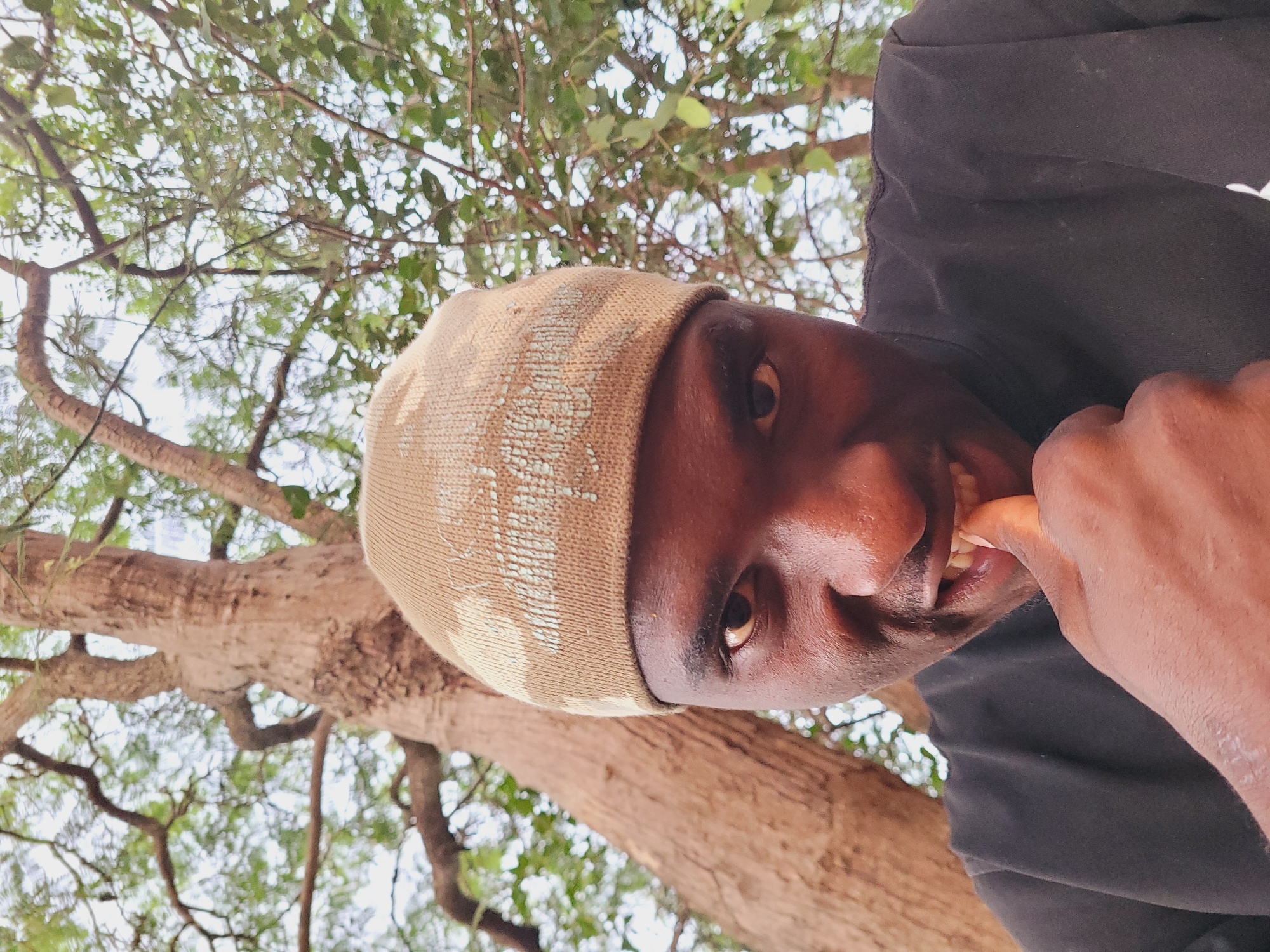In a medium-shot, a dark-skinned person wearing a tan beanie with a light pattern and a black shirt looks directly into the camera. The person's right thumb is in their mouth, and their teeth are visible. The background features a large tree with a rough brown trunk and green foliage overhead, suggesting an outdoor setting in Bukuru, Nigeria. The lighting is natural, casting subtle shadows on the face.