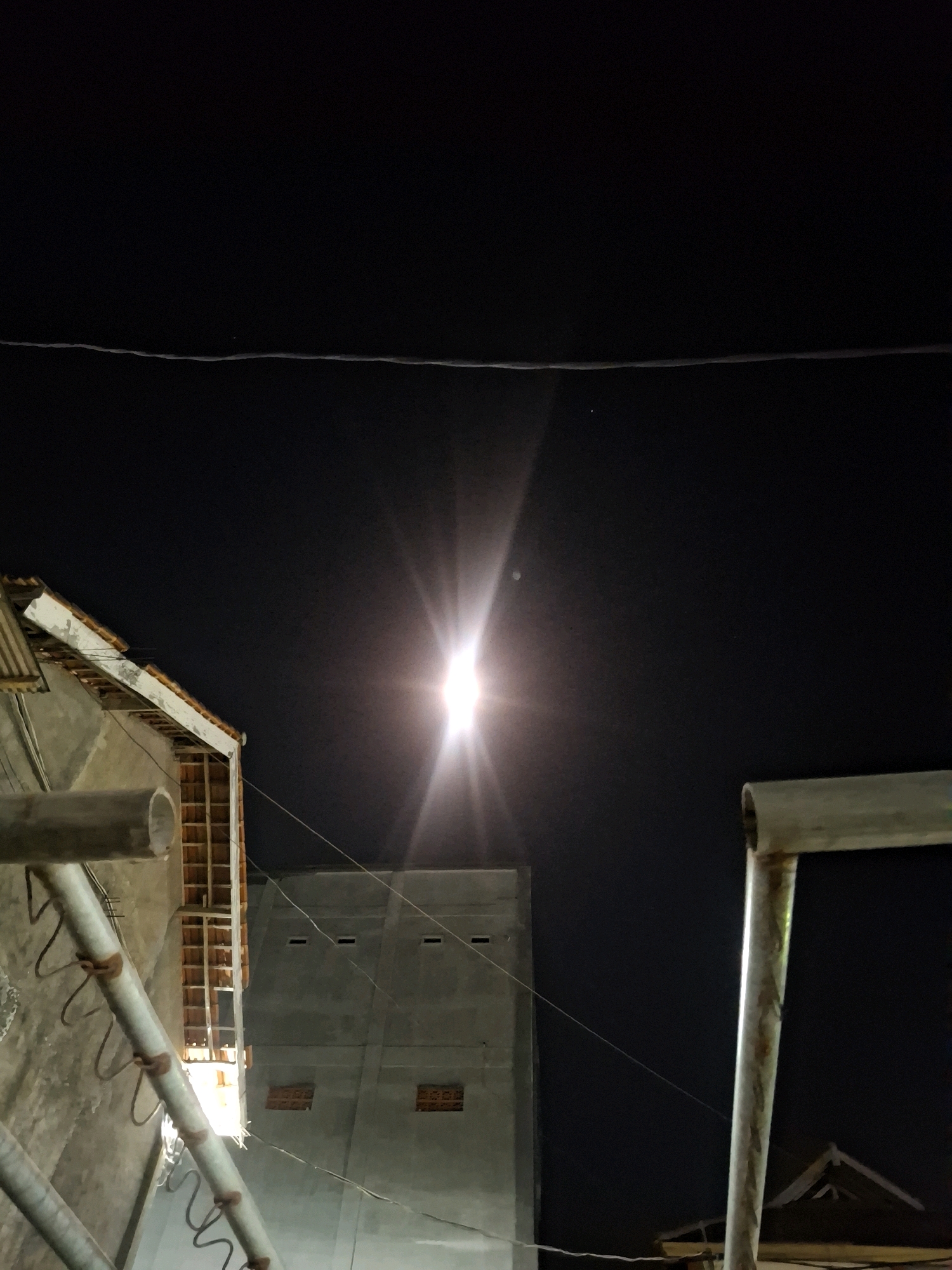 The image depicts a nighttime scene in Potoan Laok, Indonesia, dominated by a bright, lens-flared light source, likely the moon or a strong artificial light, positioned centrally in the upper half of the frame. The sky is a deep black, with a faint horizontal line, possibly a power line, stretching across the top.
In the lower left, a portion of a building with a tiled roof and concrete walls is visible. A metal pipe runs diagonally across the foreground from the bottom left, with a few hook-like fixtures attached. To the right of this pipe, another, taller building with a flat, grey concrete facade and visible windows is present. Another metal structure, a pole or scaffolding, is in the lower right foreground, also appearing metallic and somewhat weathered. The overall impression is an urban or semi-urban environment at night, with structures and infrastructure under a dark sky illuminated by a powerful light source. There are no people or discernible actions in the image.
