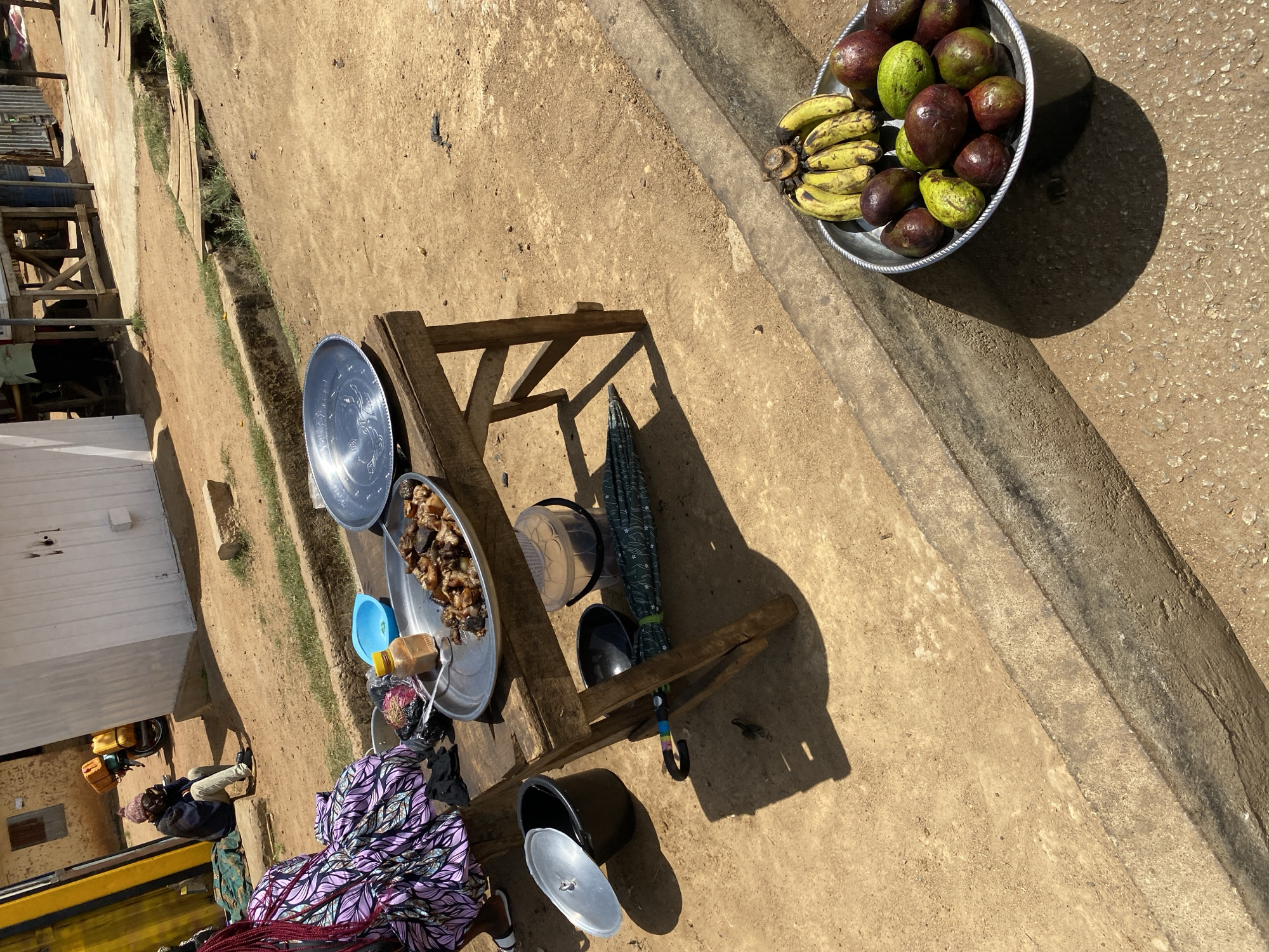An outdoor market scene, possibly in Bukuru, Nigeria, shows a wooden table with multiple silver platters holding prepared food. A blue cup and yellow container are nearby. Under the table, a black pot with a lid and a black umbrella rest. To the right, a silver bowl is full of bananas and avocados. A person wearing a dress patterned with purple leaves stands at the table. Another person sits behind a building, to the left. The ground is dirt and concrete.