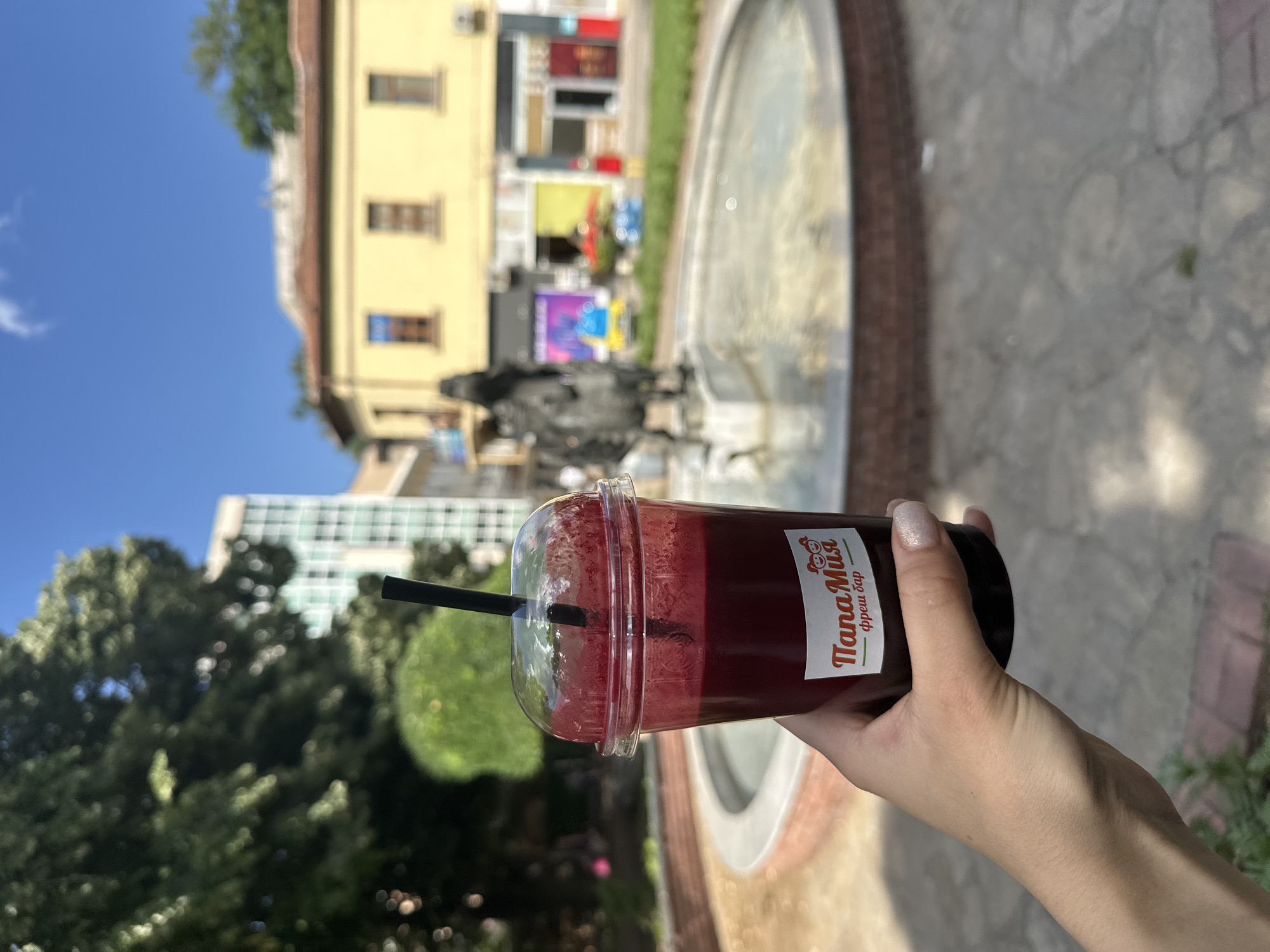 A person's hand, with neatly manicured nails, holds a clear plastic cup filled with a dark red liquid, likely a juice or smoothie, topped with a clear plastic lid and a black straw. A sticker on the cup features a logo with two cartoon bears and text that reads "ПапаМия" and "фреш бар". The background is blurred but suggests an outdoor urban setting with a fountain in the foreground, a statue within the fountain, and buildings behind it. The architecture of the buildings, particularly the yellow one with arched windows and a red tile roof, is characteristic of older European city centers. The presence of a modern glass-fronted building to the left of the yellow building indicates a mix of architectural styles. The sky is clear and blue. This scene is located in the central square of Haskovo, Bulgaria, specifically near the fountain and the iconic building of the former Municipal Bank, which now houses shops and cafes.