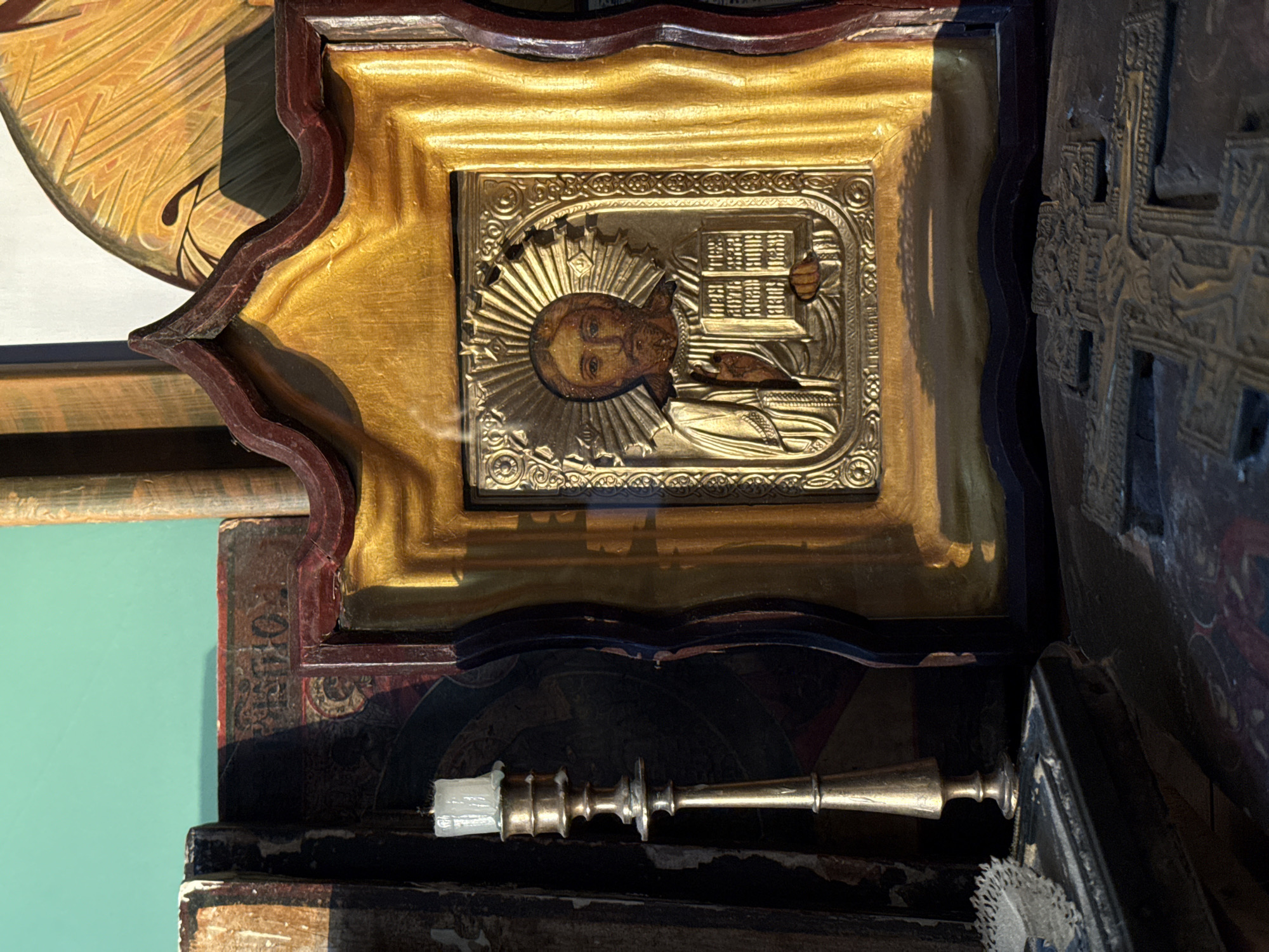 The image depicts an icon of Jesus Christ in a gilded frame, illuminated by natural light casting shadows. To the left, a brass candlestick holds an unlit white candle. The background includes aged books and religious artifacts, suggesting a domestic or spiritual setting. The scene is dimly lit with warm tones, highlighting the texture and metallic sheen of the objects. The overall atmosphere is one of solemnity and contemplation. It is impossible to determine the location within Votkinsk, Russia, from the visual information provided.