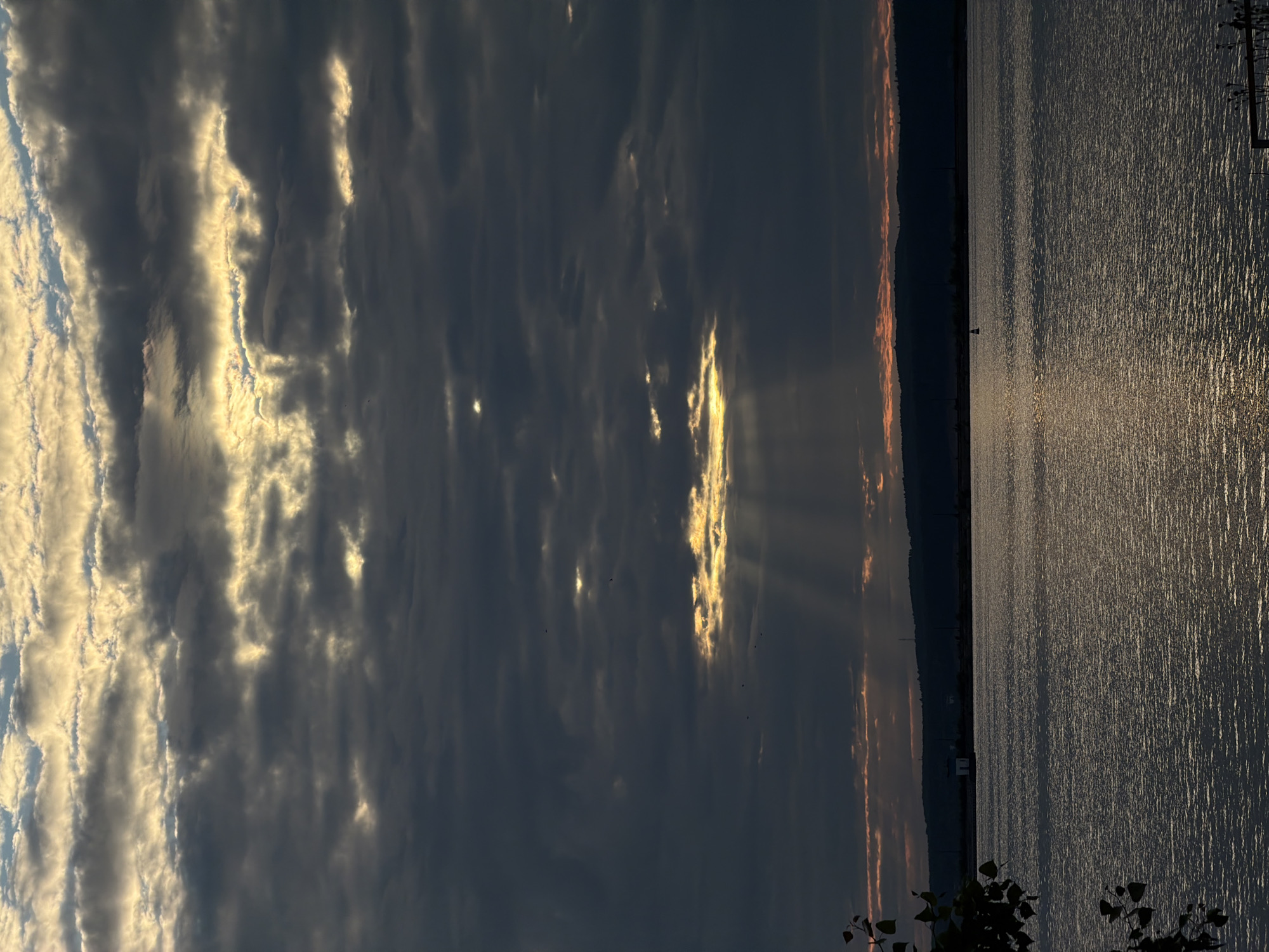 The image captures a dramatic sky over a body of water, likely the Kama River in Chaikovsky, Russia. The sky is filled with dark, ominous clouds that are broken by shafts of golden sunlight, creating a striking contrast. These sunbeams pierce through the clouds, illuminating sections of the sky and casting a warm glow on the water's surface. In the distance, a dark, silhouetted treeline forms the horizon, with hints of a faint orange glow suggesting the proximity of sunset or sunrise. On the shoreline, a small, white structure is visible, possibly a building or a lighthouse. A single, small buoy can be seen in the water, marking a point or a channel. The water itself is calm, reflecting the dramatic play of light and shadow from the sky. The foreground is partially obscured by the dark leaves of a tree on the left, adding depth to the composition. The overall mood is serene yet powerful, showcasing the raw beauty of nature.