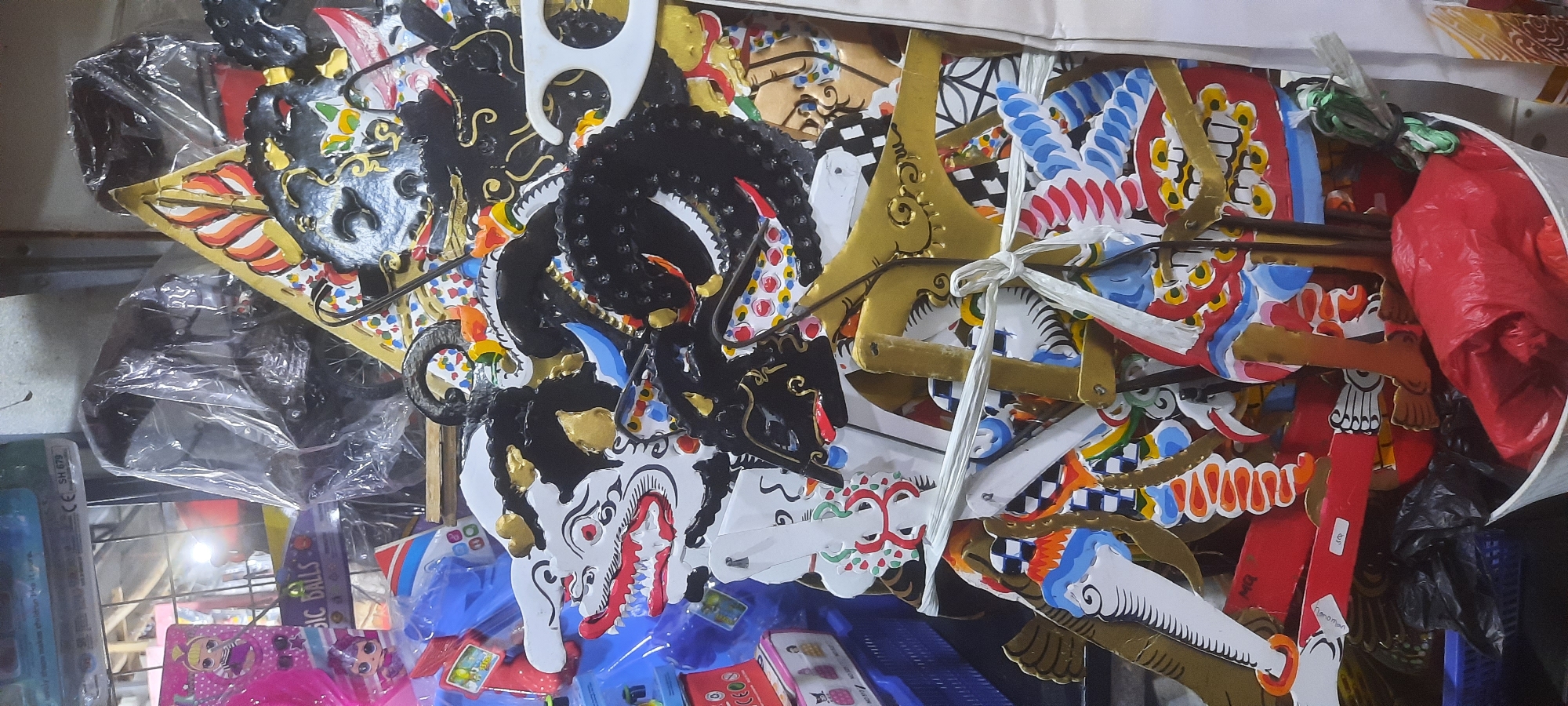 The image depicts a close-up view of a collection of Wayang Kulit puppets, a traditional form of Indonesian puppetry, displayed in what appears to be a shop or market stall. The puppets are made of intricately painted leather and are characterized by their vibrant colors and detailed designs, featuring mythical figures with exaggerated features such as sharp teeth, elaborate headdresses, and expressive eyes.
Several puppets are visible, including figures with white, black, and gold coloration. One prominent puppet on the left has a white face with red eyes and a red mouth displaying sharp teeth, a black mane, and gold accents on its headdress. To its right, another puppet has a predominantly black face with intricate gold detailing and a coiled headdress. Other puppets, partially obscured, display colorful patterns, including checkered designs, and red and blue embellishments.
The puppets are arranged in a vertical stack, with some appearing to be held together by white string. Wooden rods, used to manipulate the puppets, are visible protruding from some of them. In the background, glimpses of other items are visible, including what looks like plastic packaging, a blue plastic crate, and possibly other toys or merchandise. There are also red and black plastic bags at the bottom of the frame, suggesting the puppets are either stored or displayed in a retail environment.
The scene is brightly lit, highlighting the details of the puppets. The overall impression is one of a vibrant cultural display, likely in a location that sells traditional Indonesian crafts. The presence of "CE SH 679" on a label in the upper left corner indicates a product certification, suggesting that these items are for sale. While the image does not explicitly state the location within Blitar, Indonesia, the style of the puppets strongly suggests they are of Javanese origin, a common art form in the region. The overall setting appears to be indoors, possibly a market or a specialized shop selling cultural artifacts.