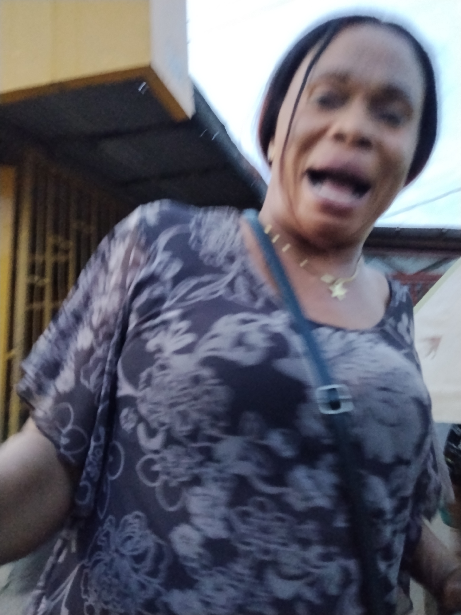 A woman wearing a floral-patterned top, a gold necklace with a star pendant, and a dark-colored crossbody bag is speaking. The scene appears to be outdoors in the city of Ura Nta Umuarandu, Nigeria, based on the prompt. There are buildings in the background.