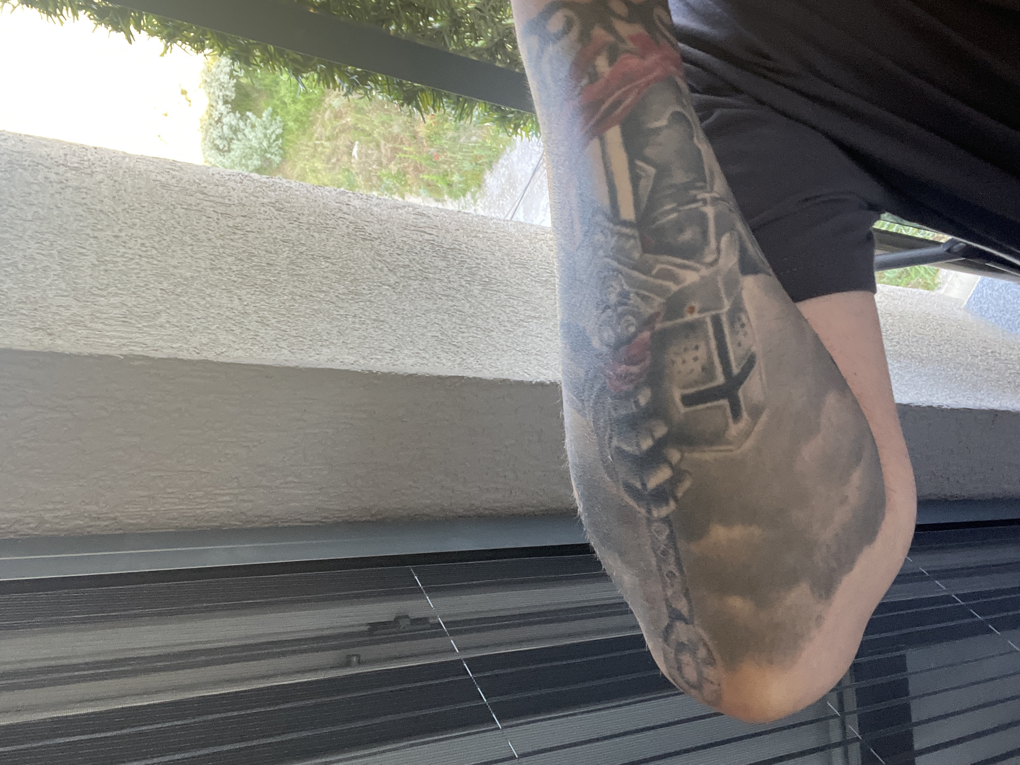 The image shows a person's tattooed arm. The tattoo appears to be a complex design with various images and symbols. The arm is bent at the elbow, and the person is wearing a dark t-shirt. The background is out of focus but appears to be an outdoor setting with a textured wall and some greenery. There is no visible indication of the image's location in Varna, Bulgaria, or any specific action or interaction taking place.