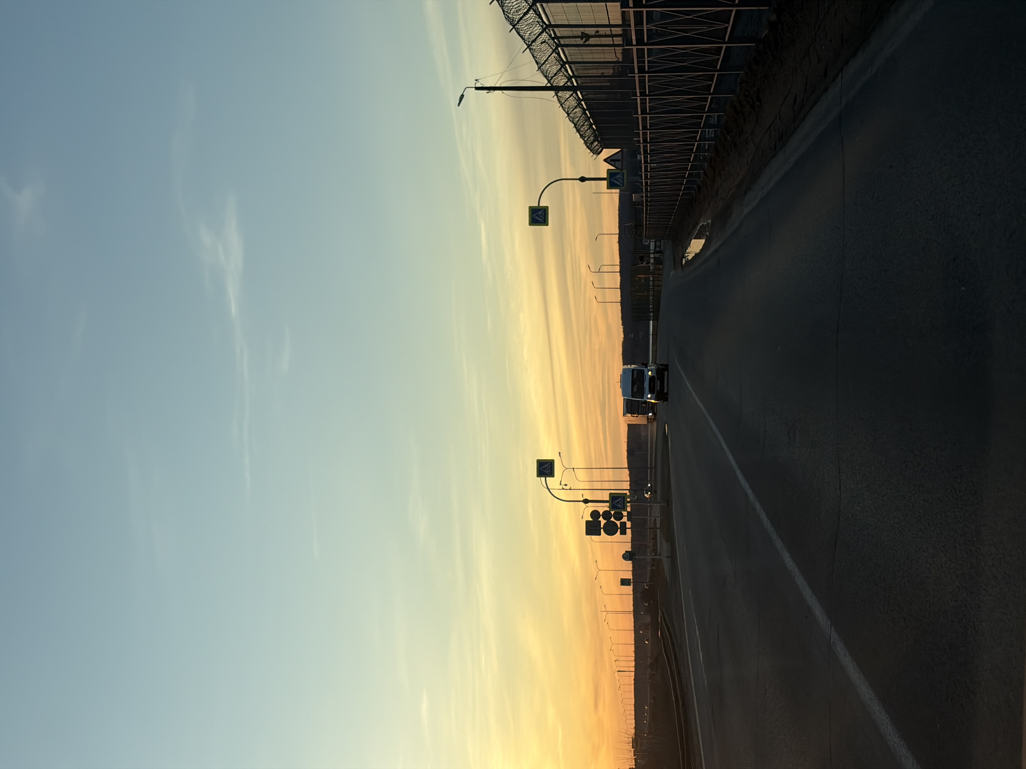 The image shows a road in Chaikovsky, Russia, facing away from the setting sun. The sky is pale blue transitioning to orange near the horizon. Along the right side of the road is a tall, dark fence. Streetlights line both sides of the road. A vehicle travels on the road. Several road signs, including pedestrian crossing signs, are visible on the right side of the road.