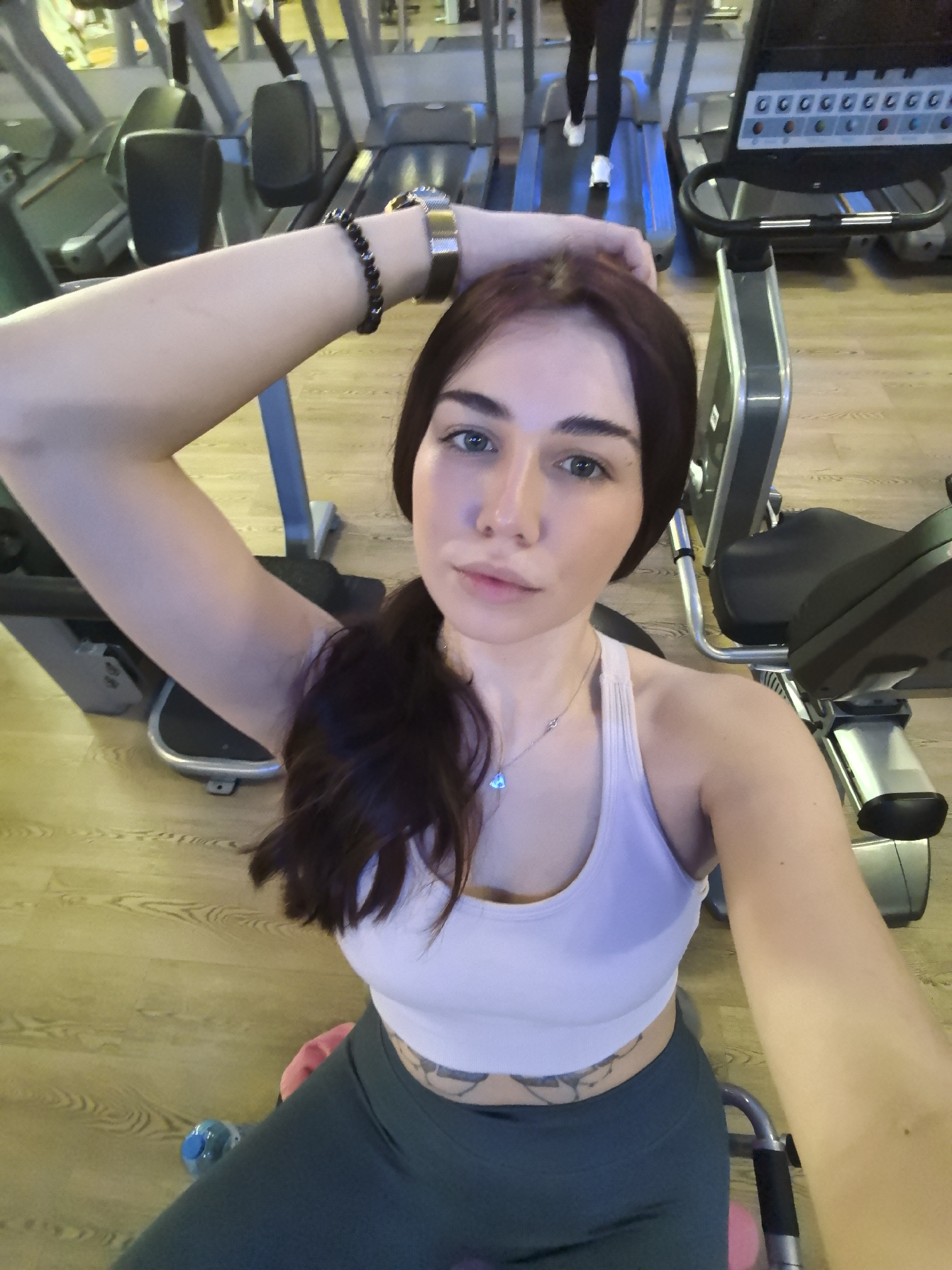 Here is a forensic description of the image:
The image shows a young woman in athletic attire taking a selfie in a gym. She is wearing a white sports bra and dark green leggings. She has a tattoo visible on her lower abdomen. Her hair is dark brown and pulled back in a low ponytail. She is wearing a watch and a bracelet on her left wrist, with her left arm raised above her head. She is wearing a necklace.
The background reveals exercise equipment, including treadmills and other weight machines. Another individual is using one of the treadmills in the distance.
Given the prompt's request for location information, it is important to acknowledge that the only location mentioned is Stara Zagora, Bulgaria. There is no information in the image to verify or confirm this location. It is only possible to describe what is visible in the image.
