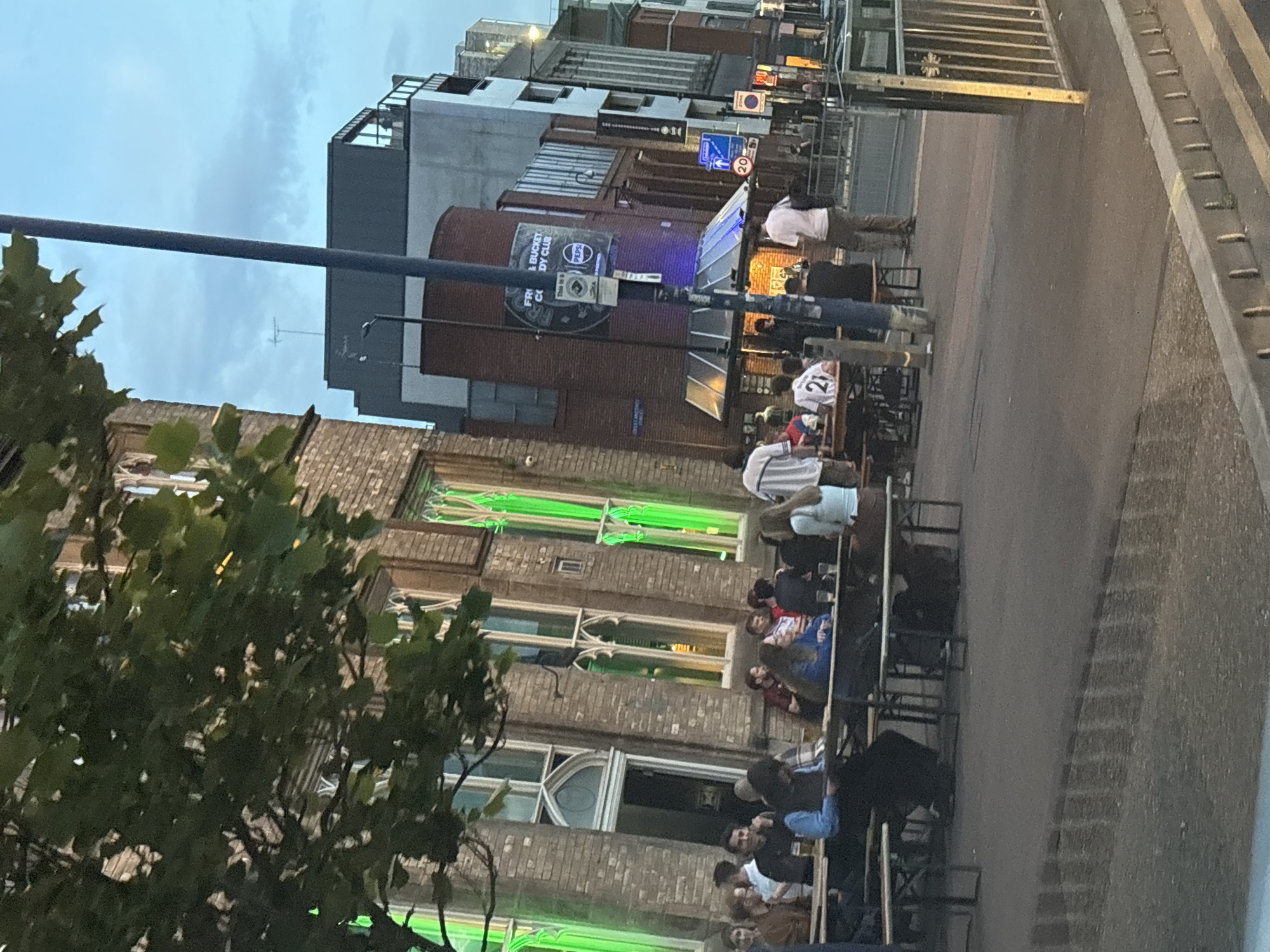 The image shows an outdoor seating area of a pub or bar in Manchester, United Kingdom. People are sitting at tables along the pavement. The architecture includes brick buildings with ornate windows highlighted by green lights, as well as more modern buildings in the background. There is a sign for "Frog & Bucket Comedy Club" with Pepsi branding visible. Street signs and traffic lights are also present. The people in the scene appear to be socializing, eating, and drinking.