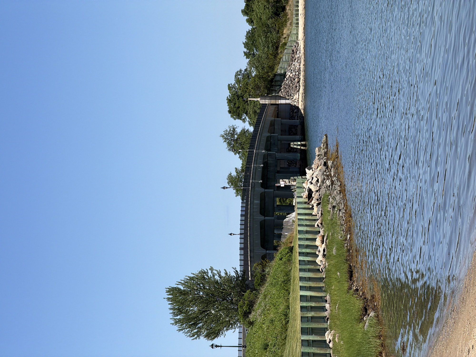 The image depicts a bright, clear day at a waterfront location, likely in the Village of Sag Harbor, United States of America. A large concrete bridge with railings and lampposts spans across a body of water. The bridge is supported by multiple concrete pillars. To the left of the bridge, a grassy hill is visible, adorned with a single, large, leafy tree and two lampposts. A wooden fence, painted green and weathered, runs along the edge of the land, separating it from the water. The shoreline is rocky with clusters of small to medium-sized rocks and patches of green grass growing near the water's edge. The water itself is a deep blue, with ripples on its surface reflecting the clear sky. On the right side of the bridge, the landscape transitions to a more densely wooded area with various trees. A sandy beach is present in the foreground on the left side of the image. There are no people or animals visible in the scene. The overall impression is one of a serene, natural landscape with a man-made structure integrated into it.