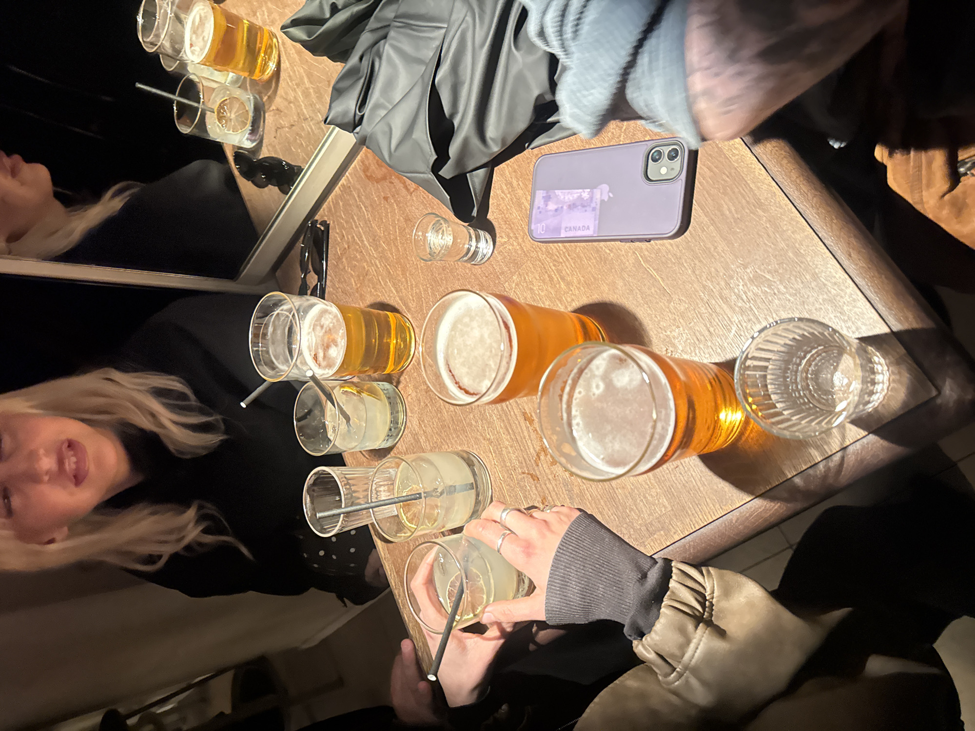 A wooden table is crowded with drinks, including two large beer glasses, three glasses with clear beverages mixed with ice and citrus wedges, and a shot glass. A purple iPhone sits on the table with a Canadian ten-dollar bill inserted under the phone case. Partial views of people are visible: a woman with blonde hair and a person holding a glass with a straw. Other items on the table include a jacket, a gray sweater, and sunglasses near a mirror that reflects the scene. The scene is dimly lit, suggesting an indoor setting such as a bar or restaurant. The location of Paris, France, cannot be determined from the image alone.