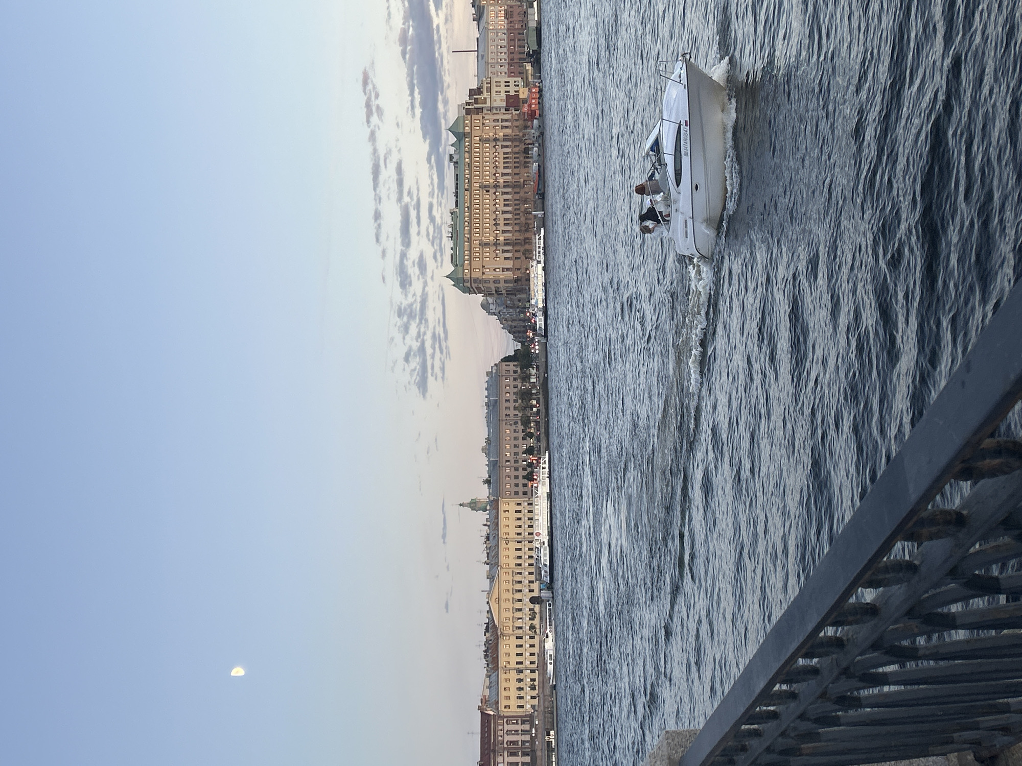 The image depicts a boat with two people aboard moving across a body of water. The boat is white and has a sleek design, with a wake trailing behind it. The water is dark and choppy, suggesting a windy day or a fast-moving boat. In the background, a city skyline is visible, characterized by numerous buildings with a uniform architectural style. The buildings appear to be old and ornate, with many windows and pitched roofs. The sky is clear and light blue, with a few wispy clouds. A crescent moon is visible in the upper left portion of the frame. The overall scene suggests a daytime setting, likely late afternoon or early evening, given the soft light. The presence of the city skyline indicates an urban environment, possibly a river or harbor. The mood is serene and peaceful, with the boat appearing to glide effortlessly across the water.