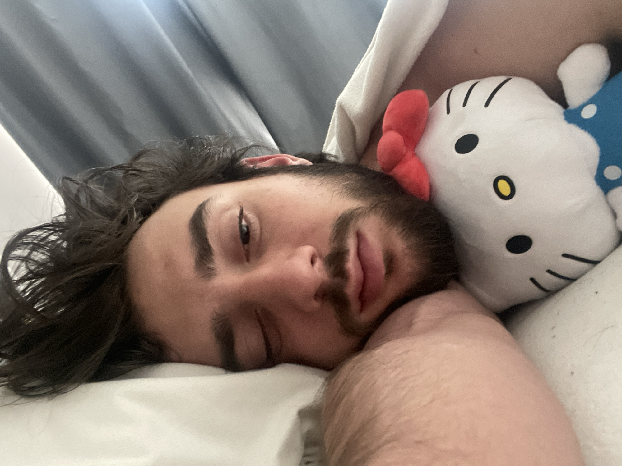 In a close-up, overhead shot, a young man lies in bed with his eyes closed, his head resting on a pillow. To his right, a plush Hello Kitty toy with a red bow rests against his cheek, its white face featuring black eyes, a yellow nose, and short black lines for whiskers. The man has dark, wavy hair, and a beard and mustache. He is wearing a blue shirt with white polka dots, and a white blanket is draped over him. The scene appears to be indoors, with gray curtains visible in the background. The lighting suggests it is daytime. The man's expression is relaxed, possibly indicating he is sleeping or resting. The overall mood is peaceful and intimate. The location context is Huixquilucan de Degollado, Mexico. There is no visible text in the image.