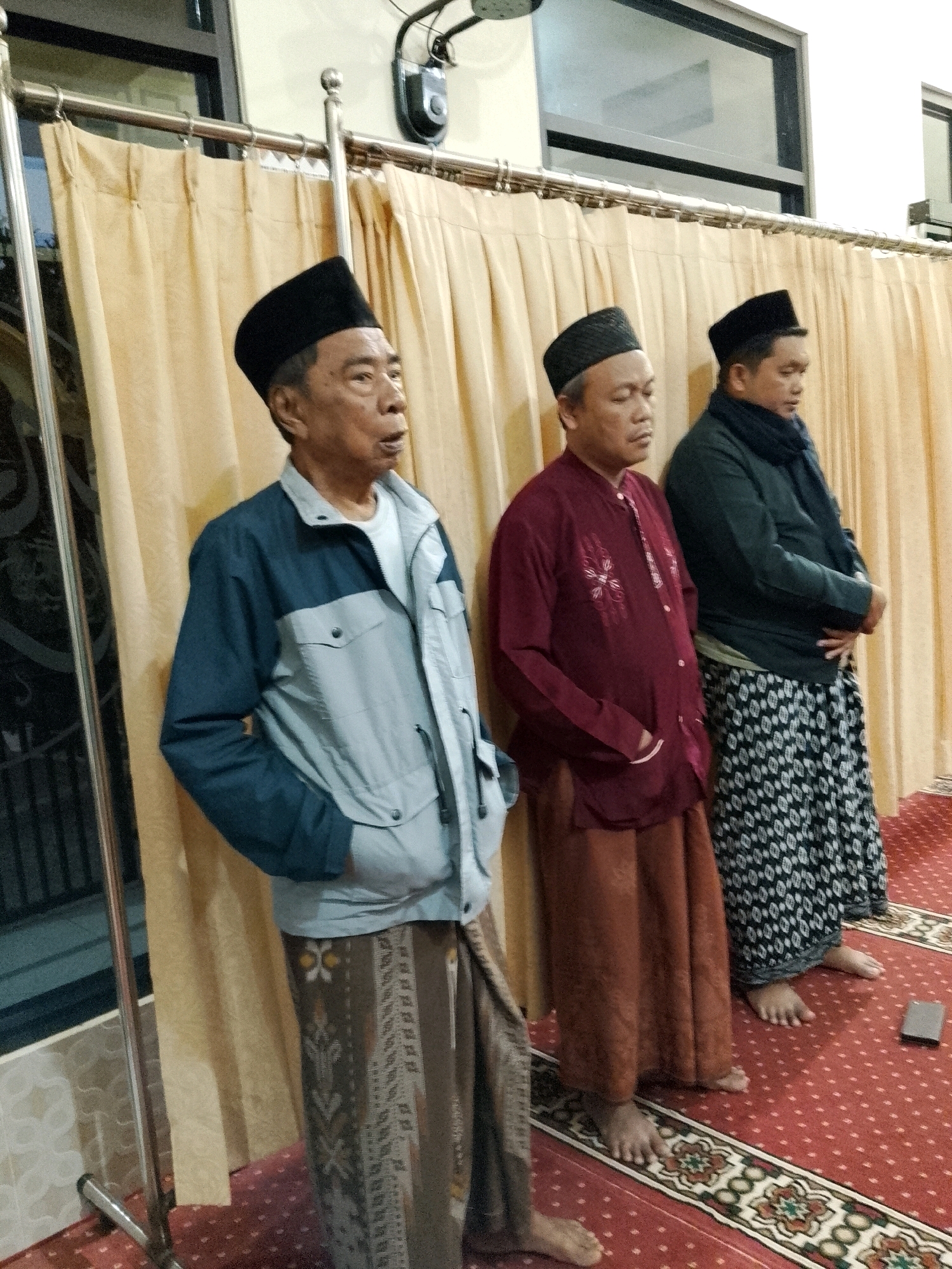 The image depicts three Muslim men standing in what appears to be a prayer hall or a communal gathering space in Rejoyoso, Indonesia. The men are dressed in traditional attire, including peci (caps) and sarongs. The man on the left wears a light blue and dark blue jacket over a white shirt and a patterned sarong. The middle man is in a maroon shirt with embroidered details and a brown sarong. The man on the right wears a dark green jacket, a scarf, and a patterned sarong. They are standing on a red carpet with intricate patterns. Behind them, a light yellow curtain hangs, partially obscuring a window or partition. A metal pole with a hook is visible on the left, possibly part of a curtain railing or partition. The lighting suggests an indoor environment, possibly during daytime or with artificial lighting. The men appear to be in a moment of reflection or prayer, with their heads slightly bowed and their hands clasped or at their sides.