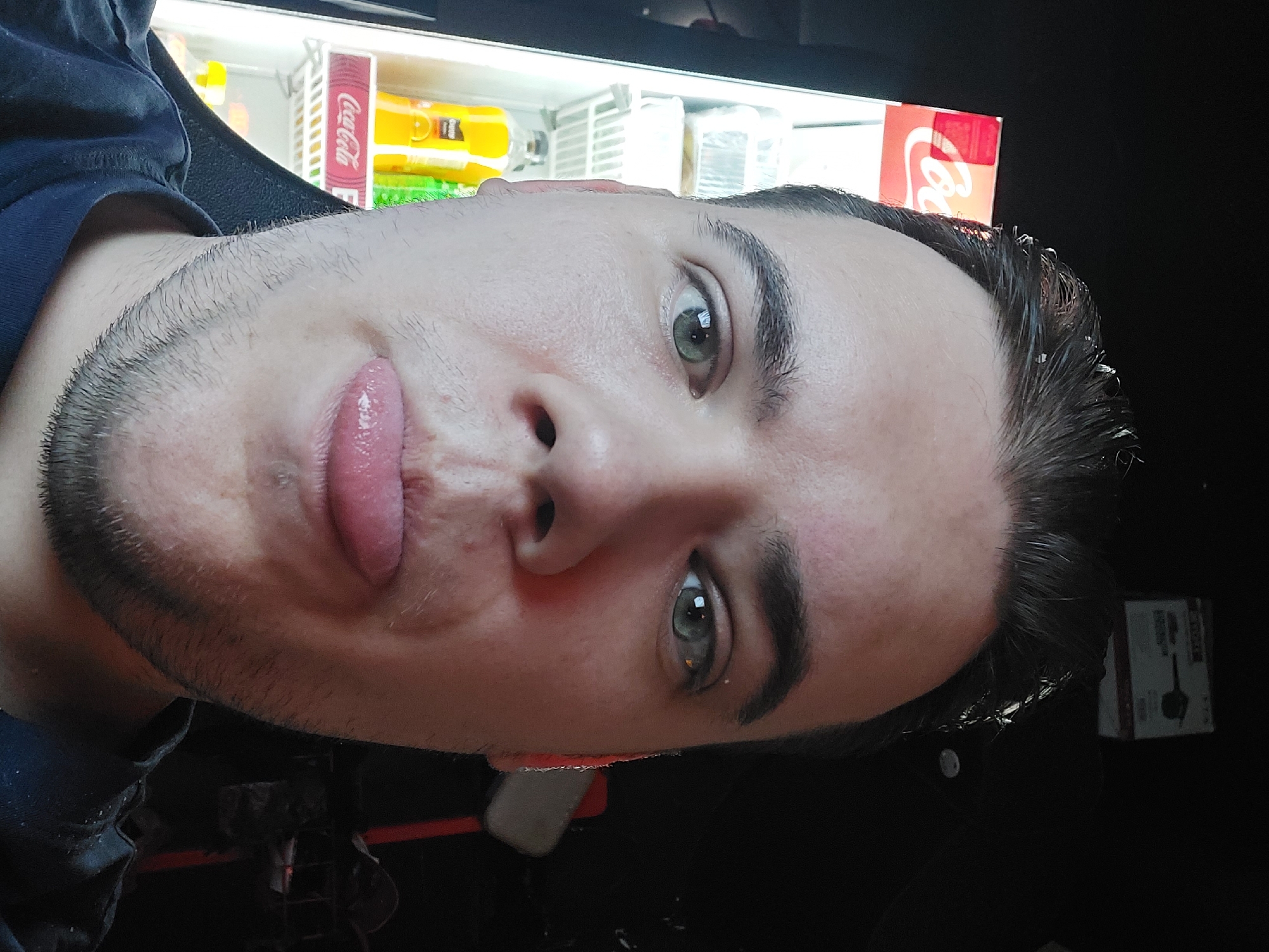 A close-up selfie of a light-skinned male with dark hair and stubble is the primary subject. He is sticking out his tongue. Behind him is a drink refrigerator with Coca-Cola branding. The location is undetermined, and the context is insufficient to specify Tashkent, Uzbekistan.