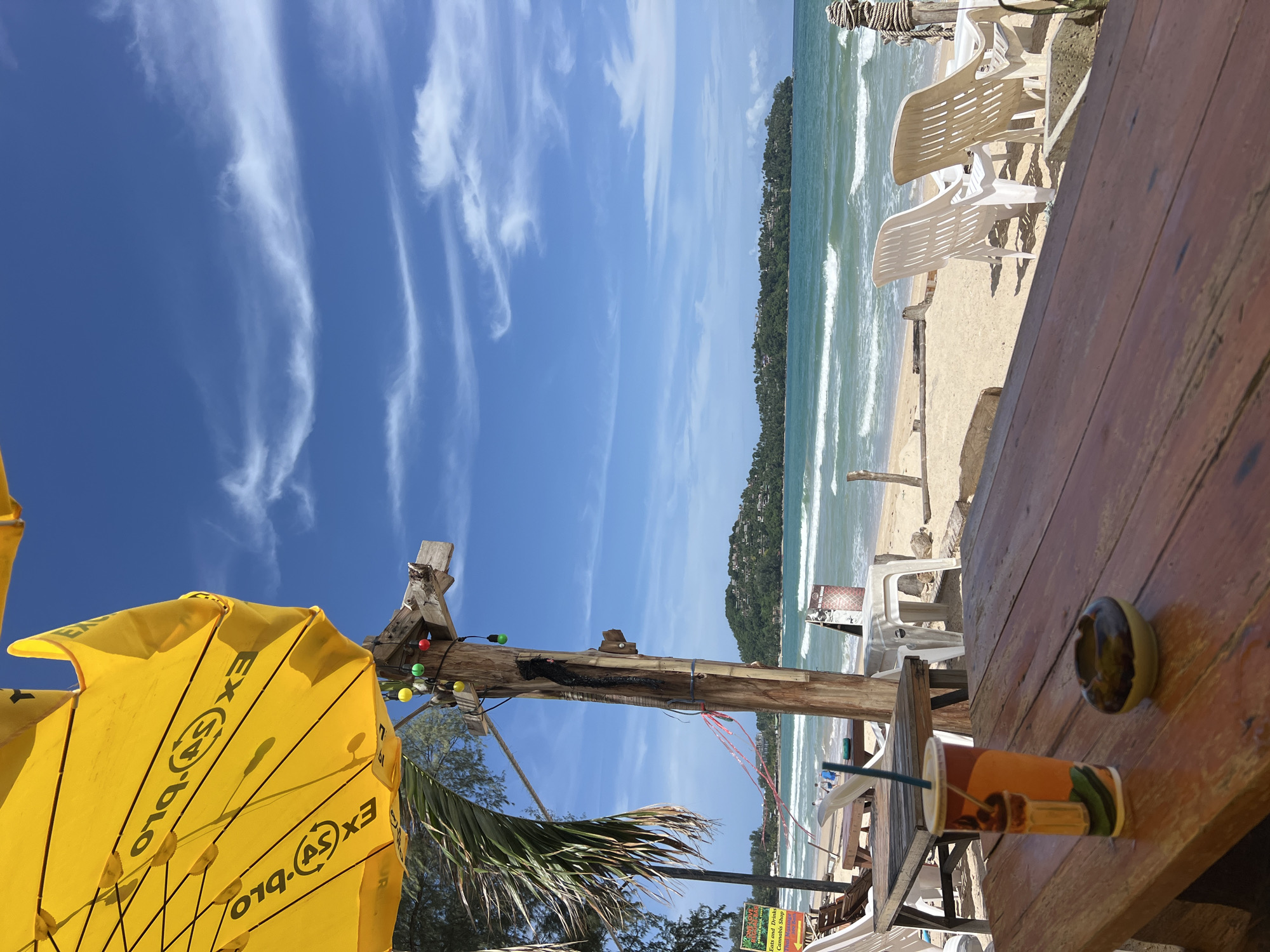 The image depicts a beach scene on a sunny day. In the foreground, a wooden table holds a colorful beverage cup with a straw and a small ashtray. Behind the table, a wooden structure with colorful lights and a palm tree are visible. The beach itself is sandy with several white plastic lounge chairs. The ocean is turquoise with gentle waves, and a distant green, tree-covered island is visible on the horizon under a bright blue sky with wispy white clouds. A large yellow umbrella is partially visible in the upper left corner of the image. There are no people clearly visible in the immediate foreground or interacting with the objects.