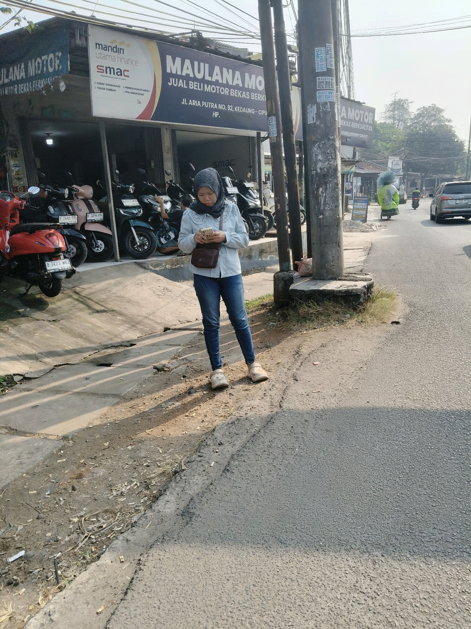 A young woman wearing a hijab and blue jeans stands on a sidewalk in front of a motorcycle dealership in South Tangerang, Indonesia. She is looking down at her phone, which she holds in her right hand. A brown crossbody bag hangs on her left shoulder. The dealership, "MAULANA MOTOR," has a banner advertising "JUAL BELI MOTOR BEKAS BERKUALITAS" (Sale and Purchase of Quality Used Motorcycles). Several motorcycles are parked in front of the shop. The sidewalk is cracked and has debris scattered on it. A paved road runs alongside the sidewalk, with traffic visible in the background. The scene is illuminated by bright sunlight, casting shadows on the ground.