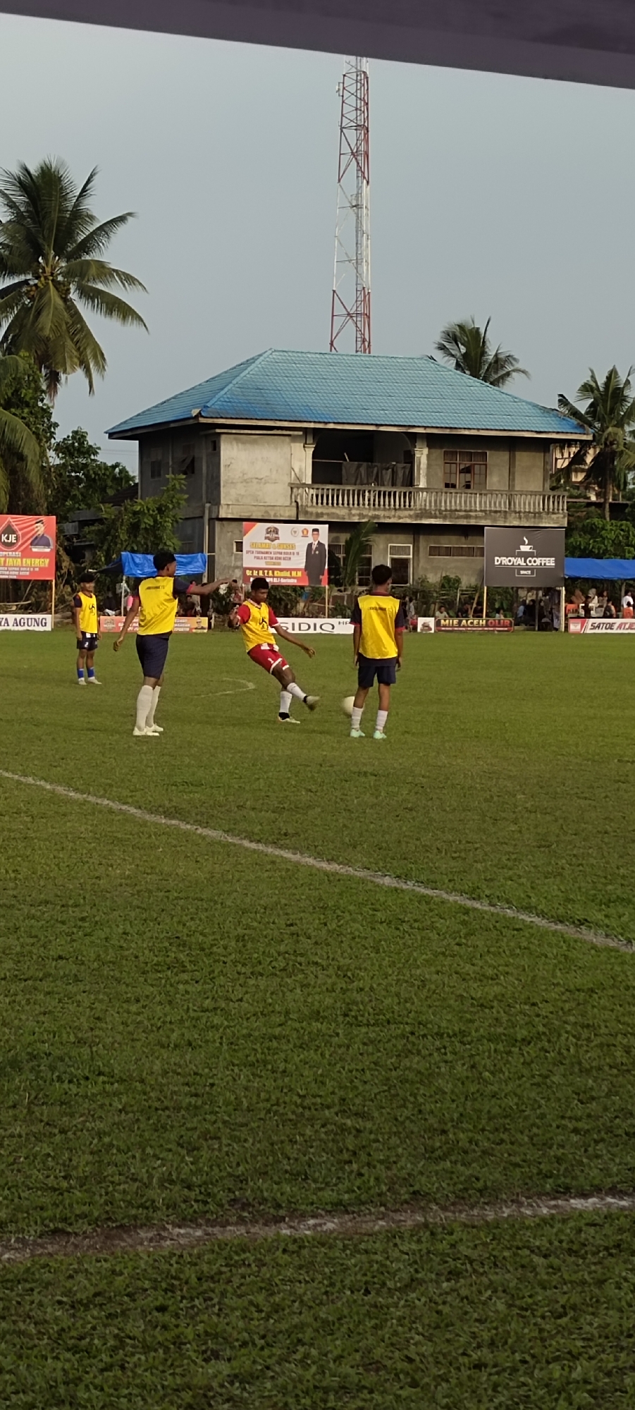 In Kilometer Delapan, Indonesia, a soccer match is underway on a well-maintained grassy field. Four young male players, all wearing yellow vests over their athletic uniforms, are visible on the field. One player in a red and yellow uniform is captured mid-action, kicking a white soccer ball. Another player in a yellow vest and dark shorts stands to his right, observing the play, while a third player in a yellow vest and dark shorts is further to the right, also observing. A fourth player, in a yellow vest and dark shorts, stands to the far left of the frame, facing away from the camera. Behind the players, a two-story building with a blue roof stands prominently. Several banners are displayed on the building, including one with the text "KUE JAYA ENERGY" and another with "D'ROYAL COFFEE". A tall, red and white telecommunications tower rises into the pale sky in the background. Palm trees are visible on the left and right sides of the scene, suggesting a tropical or subtropical environment. The overall scene depicts a community sporting event.