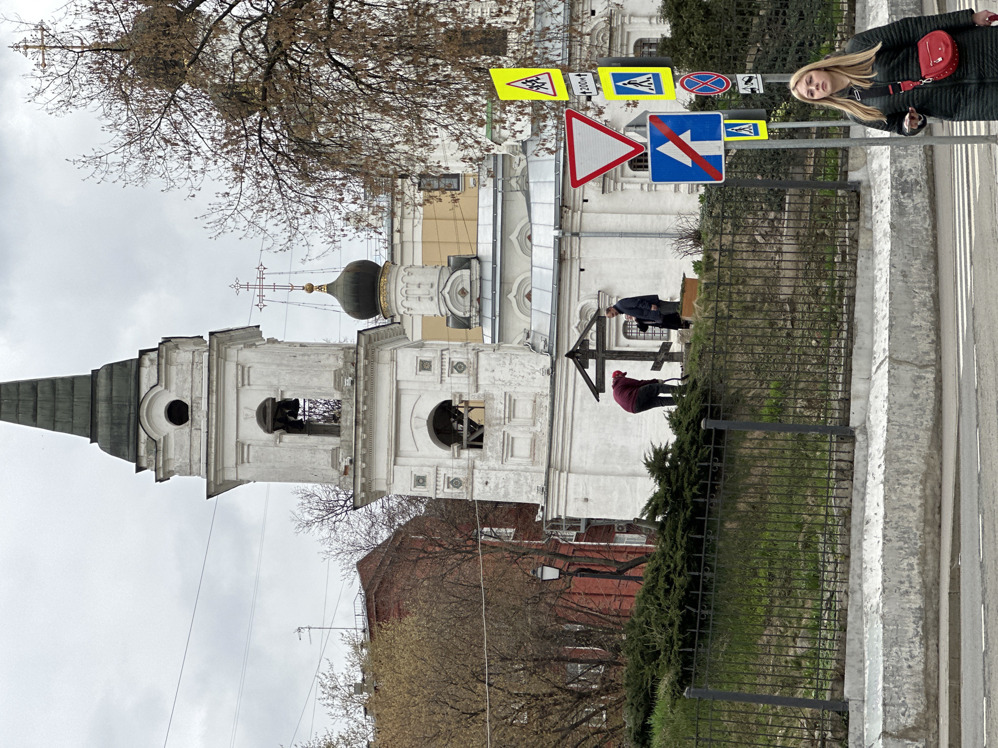 The image captures a street scene in Moscow, Russia, featuring an Orthodox church prominently in the background. The church is white with a black dome and gold accents. A bell tower is attached to the main structure. Several bare trees are scattered around the church, suggesting it may be autumn or winter.
In the foreground, a black metal fence runs along the sidewalk. On the sidewalk, a woman with long blonde hair is walking, wearing a black jacket and a red purse. Behind the fence and closer to the church, two men are standing near a dark cross or monument.
Several traffic signs are visible next to the sidewalk. The weather appears overcast with a gray sky. A red brick building is partially visible behind the church and trees.