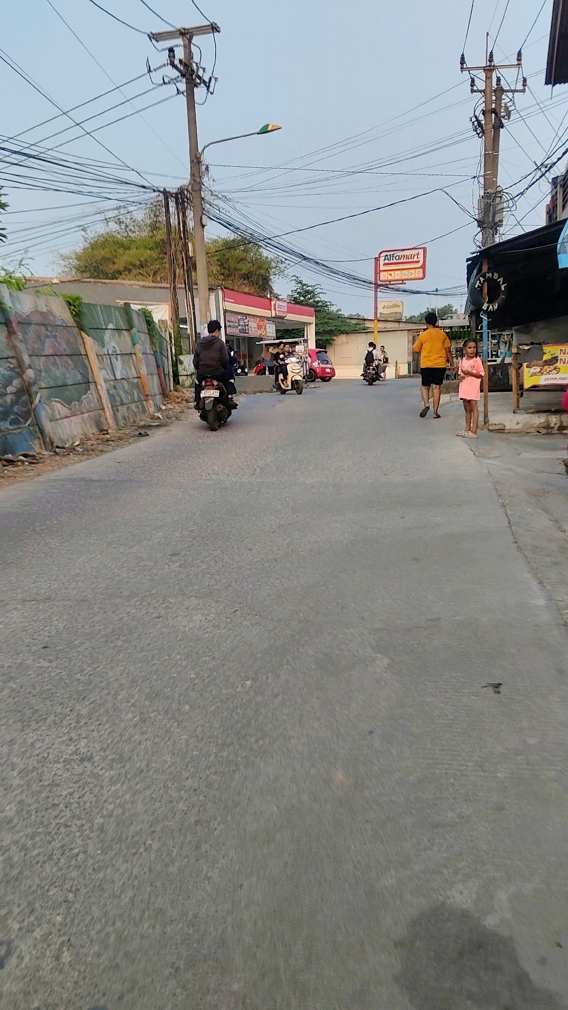 The image depicts a street scene in Mekarmukti, Indonesia, at what appears to be dusk. A paved road occupies the foreground, with a slight incline. On the left side of the road, a low wall is adorned with colorful graffiti, and beyond it, a building with a red awning, possibly a convenience store, is visible. Several people are on or near motorcycles. One person rides a motorcycle away from the viewer, while others are stopped or parked near the building. In the mid-ground, two individuals, a man in a yellow shirt and a young girl in a pink dress, are walking on the right side of the road. A sign for "Alfamart" is prominently displayed on a red and white pole. Utility poles with numerous wires crisscross the sky, and a street lamp with a multi-colored shade is mounted on one of them. The overall atmosphere is one of a typical urban street in Indonesia, with active local commerce and pedestrian and vehicular traffic.