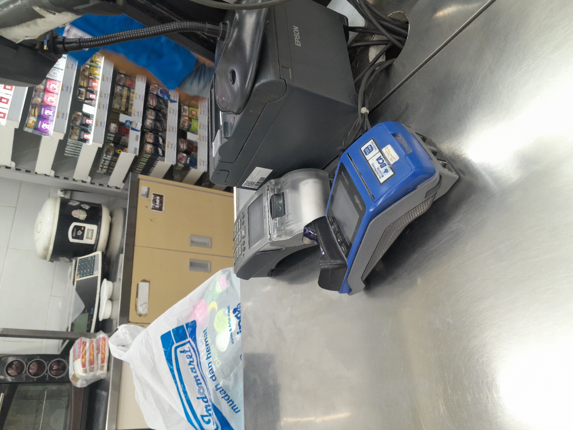 The image captures a close-up view of a retail counter in what appears to be a convenience store in Desa Ponokawan, Indonesia. The counter surface is made of stainless steel, reflecting the overhead lighting. On the counter are two electronic payment terminals, one silver and one blue, positioned in front of a dark gray receipt printer with "EPSON" visible on its side. To the left, a partially visible white plastic shopping bag with the "Indomaret" logo is present, suggesting a purchase is being processed or has recently been made. In the background, shelves stocked with various products, including what seem to be food items and personal care products, are visible. An employee wearing a blue shirt and denim shorts is partially visible behind the counter, indicating a service transaction. The overall scene depicts a typical checkout area of a retail establishment.