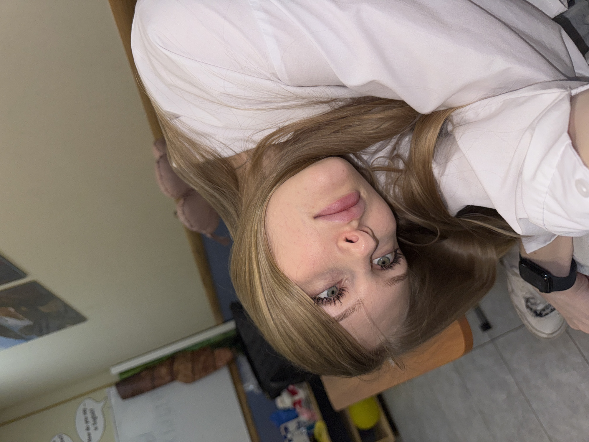The image shows a young woman with light blonde hair and green eyes. She is wearing a white collared shirt and has a black smartwatch on her left wrist. Her face is tilted upwards towards the camera. The background appears to be a room with light-colored walls and a whiteboard with writing on it. There is also a chair and various objects on the floor, including what seems to be cleaning supplies. The location is indicated by the presence of Russian language on the whiteboard. The location is in Perm, Russia.