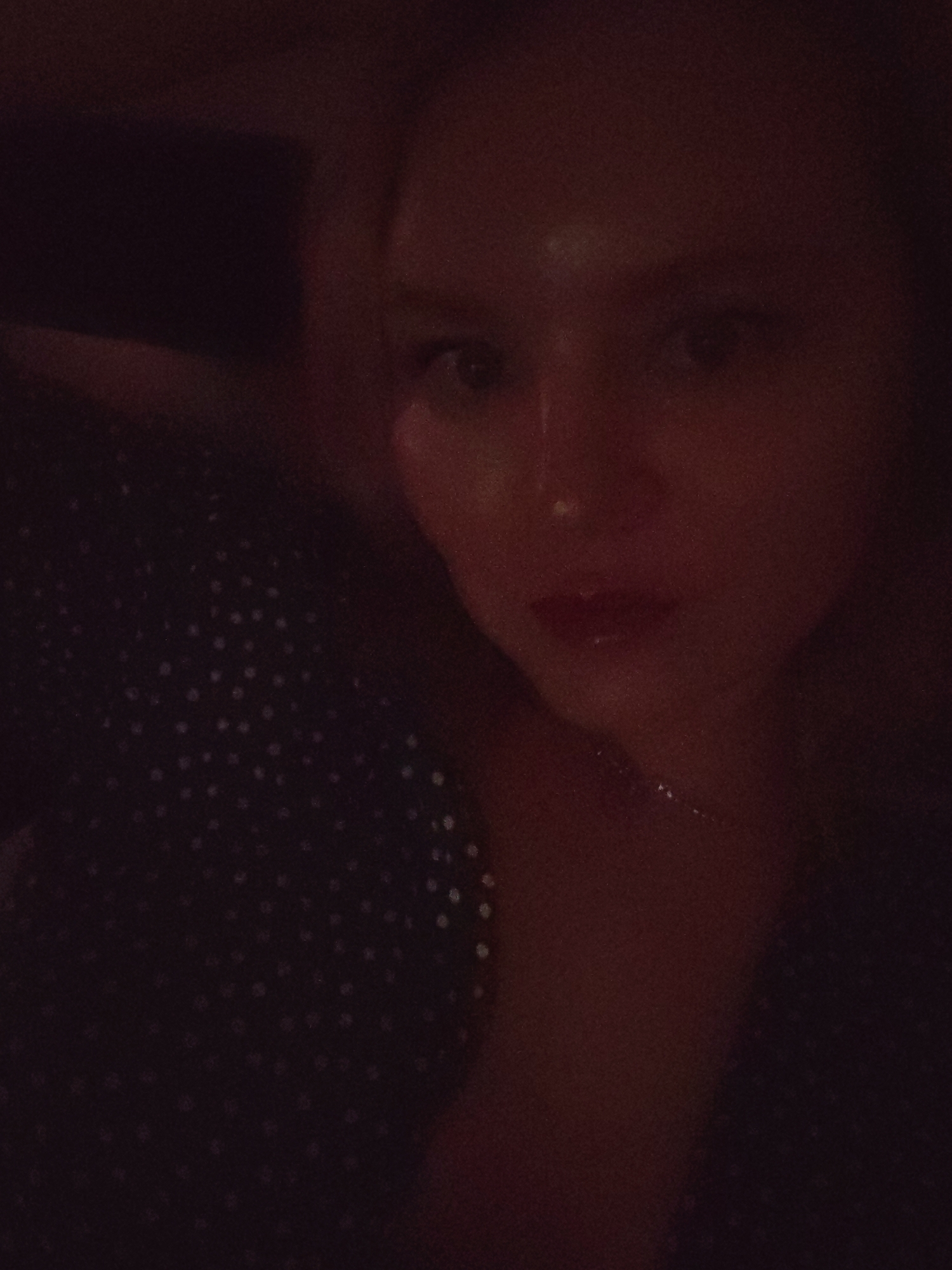 A woman with fair skin and blonde hair is posing for a selfie in low light. She is wearing a dark, patterned top. A necklace is faintly visible around her neck. The location is not discernible from the image.