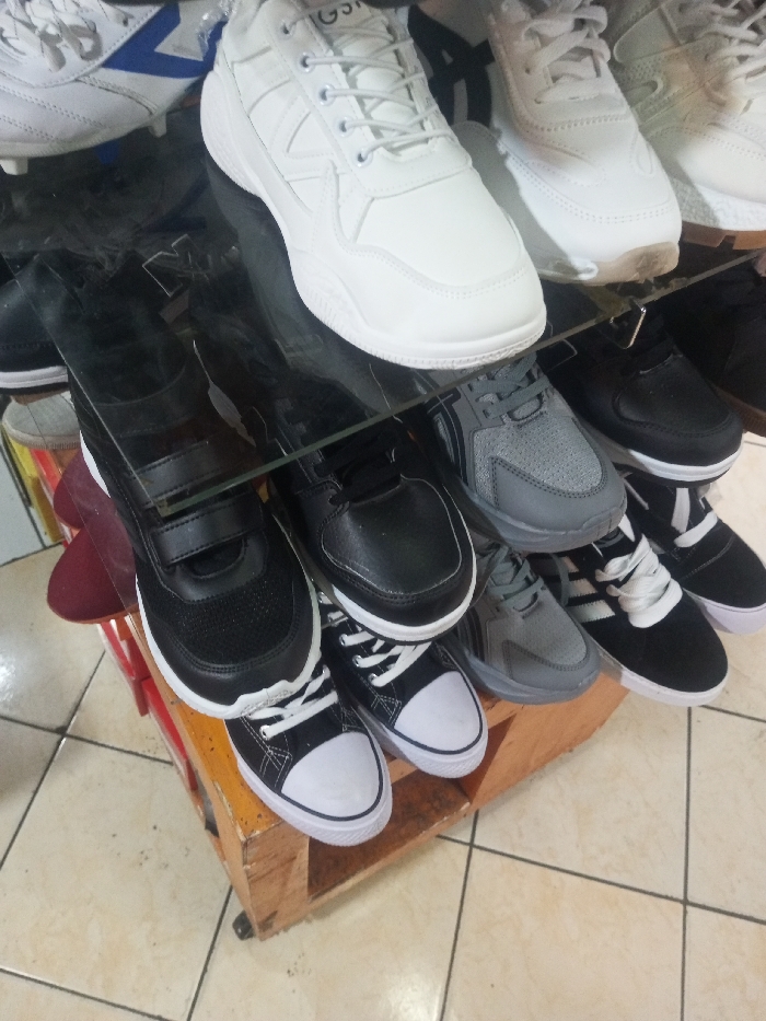 A variety of shoes are displayed on a tiered shelf. The top shelf contains white sneakers and white cleats. The second shelf features gray and black sneakers. The bottom shelf holds black sneakers, some with Velcro straps, alongside black and white canvas shoes. The shoes are arranged in neat rows, showcasing different styles and colors. The shelf is made of wood.