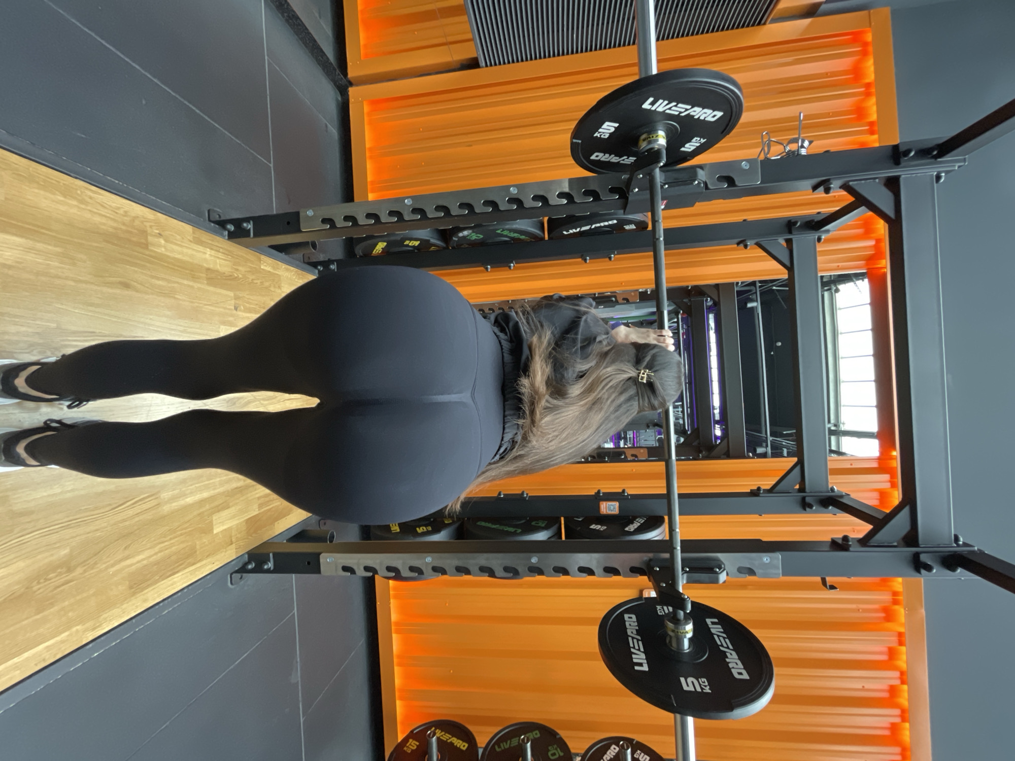 Here is a detailed description of the image:
**Scene:** An indoor gym setting, likely in Kazan, Russia based on the potential for Russian text appearing on equipment and the overall style of the facility, although this needs verification.
**Subject:** A woman wearing dark leggings is positioned beneath a barbell rack. She is facing away from the camera, allowing a clear view of her attire and posture. The focus is on her lower body and the equipment around her.
**Objects:**
* **Barbell Rack:** A sturdy, black metal rack with orange accents is present, designed to hold a barbell at different heights. It appears to be equipped with additional weight storage options on the sides.
* **Barbell:** A steel barbell runs vertically through the center of the frame.
* **Weight Plates:** Circular black weight plates, labeled with "LIVEPRO" and weight denominations such as "5 KG", are affixed to the barbell. Additional weight plates are neatly stacked on storage pegs along the rack.
* **Flooring:** The gym floor is a combination of light wooden flooring and a darker grey surface.
**Actions:** The woman appears to be preparing for or in the process of performing a barbell exercise, potentially squats, deadlifts, or a similar lower body workout.
**Environment:** The gym has a modern aesthetic with bright orange accents along the walls and equipment. The lighting seems bright. The gym appears clean and well-maintained.