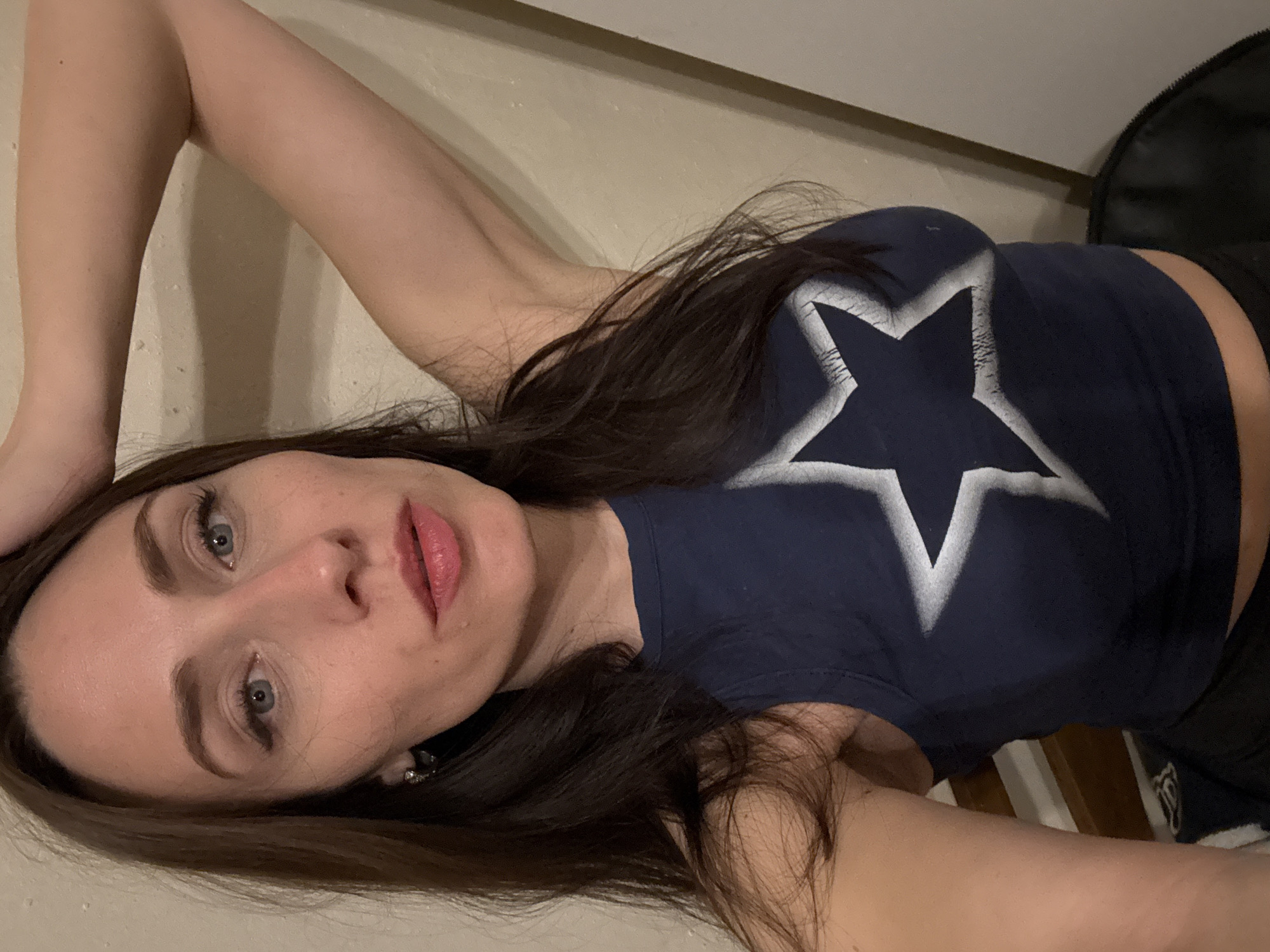 A woman is depicted in a medium close-up shot, oriented horizontally as if she is lying down and looking towards the camera. She has long, dark brown hair that spreads around her head, light blue eyes, and fair skin. Her lips are slightly parted, revealing a pinkish hue. She wears a dark blue, fitted crop top with a prominent white star design on the front. The star appears to have a slightly distressed or textured finish.
Her right arm is raised, with her hand positioned near the top of her head, and her left arm is partially visible beneath her. Her gaze is direct and neutral. The background behind her consists mainly of a light, uniform surface, likely a wall or bed linen. To the upper right, a dark, soft object, possibly a bag or jacket, is partially visible. In the lower right of the frame, there are glimpses of dark wooden elements, suggesting furniture like a bed frame or headboard. The lighting is soft and even, indicating an indoor setting.