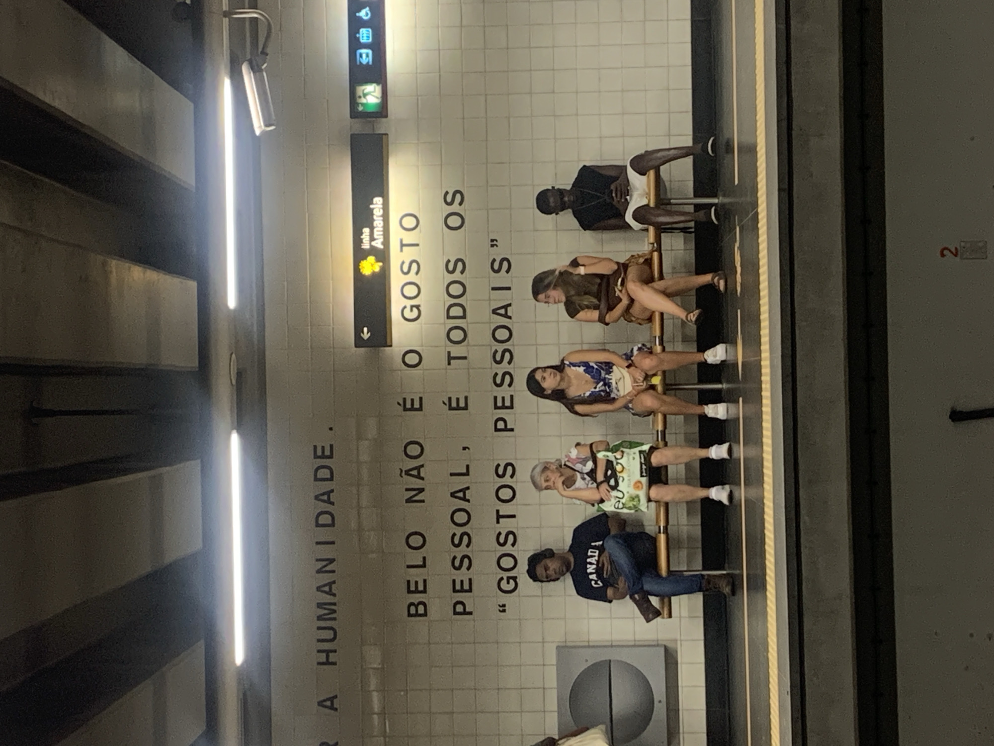 The image depicts a subway station in Lisbon, Portugal, specifically on the Yellow Line (Linha Amarela), as indicated by a sign in the upper right. The station features a tiled wall with large, dark lettering that reads, "Belo não é o gosto pessoal, é todos os 'gostos pessoais'". In the foreground, five individuals are seated on a bench, waiting. From left to right, they are a man wearing a black t-shirt with "CANADA" printed on it and jeans, a woman with gray hair wearing a floral print dress and holding a green bag, a woman in a blue and white floral dress, a woman in a brown top and a light-colored skirt, and a man in a black sleeveless shirt and white shorts. The background shows a white tiled wall, fluorescent lighting, and a security camera. The overall atmosphere is that of a typical urban public transportation hub.
