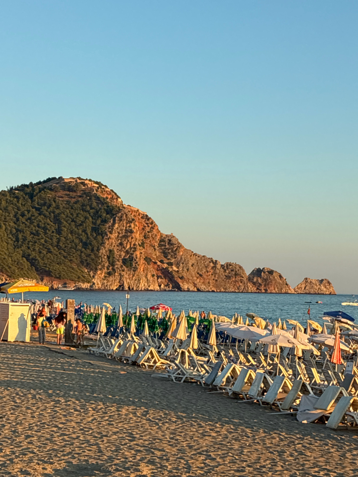The image depicts a crowded beach scene in Alanya, Turkey, with the prominent Alanya Castle complex visible on a rocky peninsula in the background. The sun is low in the sky, casting long shadows across the sand. Numerous beach chairs and umbrellas are set up in neat rows, suggesting a popular tourist destination. People are scattered throughout the beach, some walking, others relaxing. The sea is calm and blue, with a few small boats visible in the distance. The overall atmosphere is one of a bustling beach day in a picturesque Mediterranean setting.