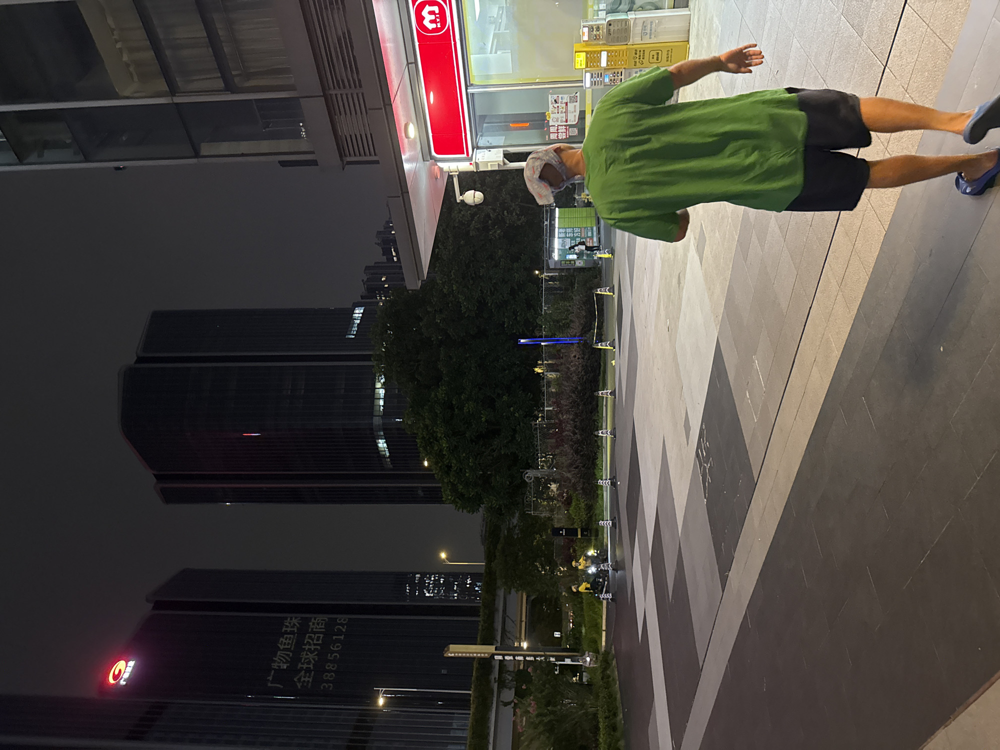 A man wearing a green t-shirt and dark shorts walks away from the camera at night on a city sidewalk in Huangpu District, China. He has a blue bandana on his head and is wearing blue slides. The sidewalk is paved with large tiles in a geometric pattern of grey, beige, and dark grey. In the background, there are modern skyscrapers with illuminated windows, and a green tree-lined area. To the right, there is a red and white convenience store with a M.V.I. logo and yellow vending machines. Two individuals on motorcycles wearing yellow uniforms are visible in the mid-ground. The overall scene is illuminated by artificial light from the buildings and store.