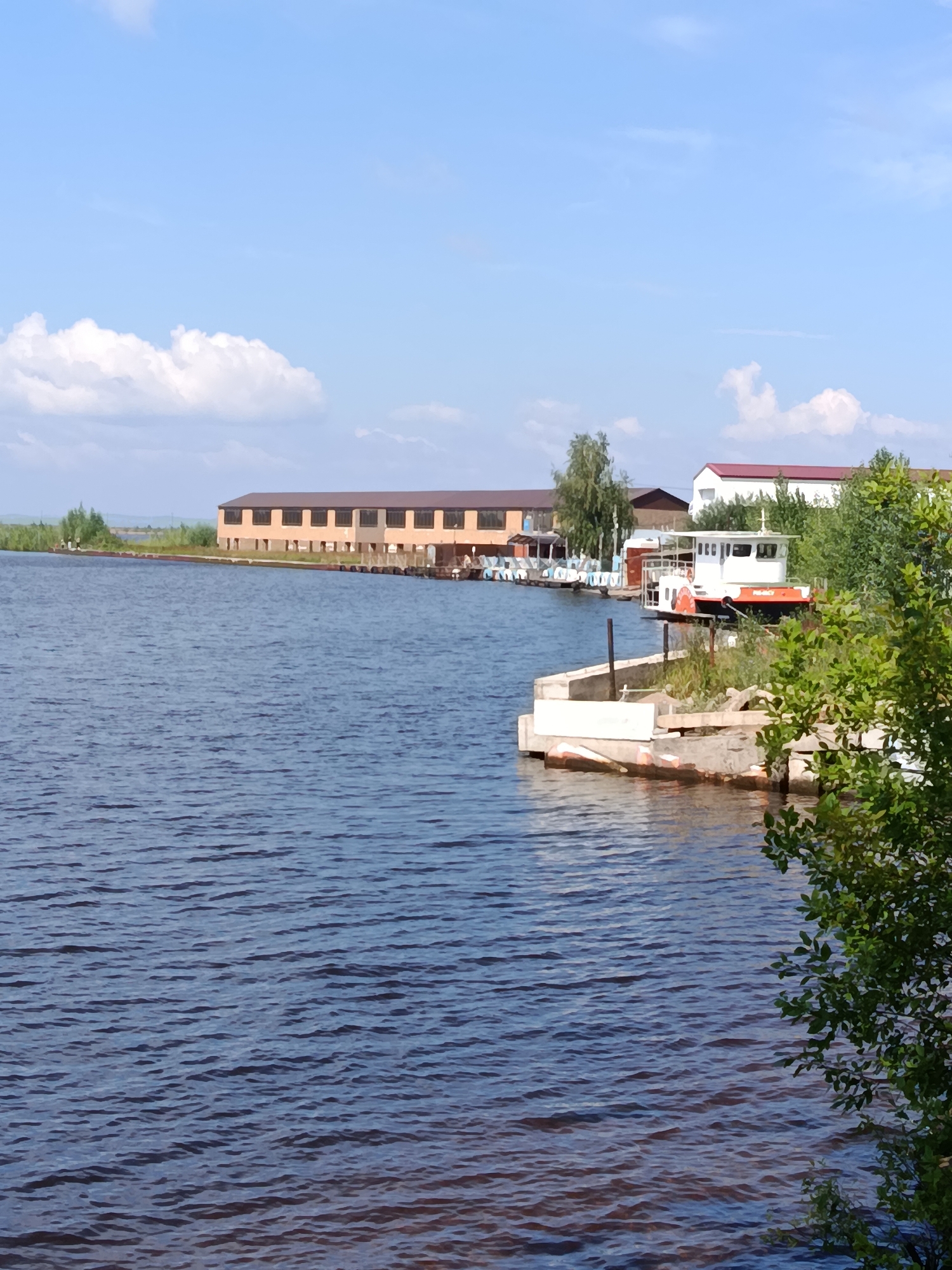 The image shows a wide body of water with a building and a boat on the shore. The water is dark blue and has small waves. In the background, there is a long, light brown building with many windows. To the right of the building, there is a white boat with an orange stripe, docked next to a concrete structure covered in plants. Green trees are visible around the boat and in the distance. The sky is light blue with a few white clouds. The scene appears to be a waterfront area, possibly a harbor or a riverside.