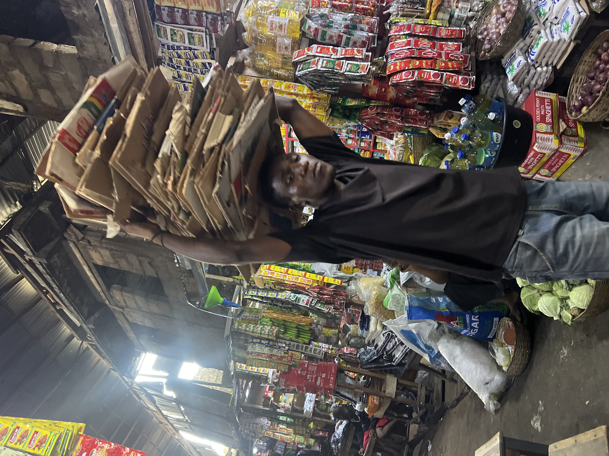 A man, likely a vendor, carries a large stack of flat cardboard boxes balanced on his head within a covered marketplace in Nkpor, Nigeria. He is wearing a dark t-shirt and blue jeans. The market stall behind him is filled with various goods including bottles of cooking oil, sacks of sugar and pasta, baskets of cabbage and onions, and numerous small packets of condiments and spices hanging from the ceiling. The marketplace appears to be constructed with corrugated metal roofing and rough brick walls, with natural light filtering in from openings. Other shoppers and vendors are visible in the background, suggesting a busy commercial environment.