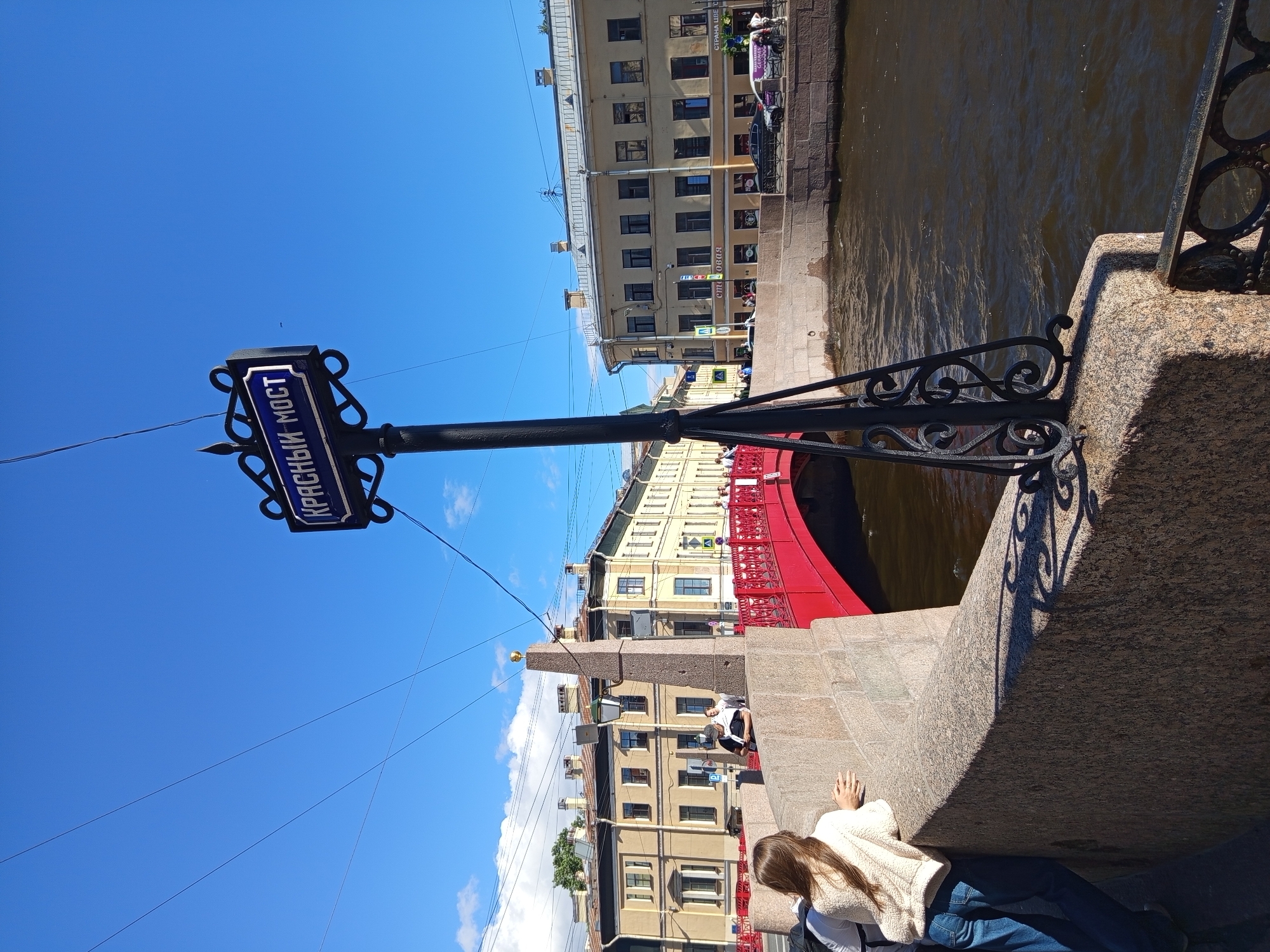 The image depicts a scene in Saint Petersburg, Russia, specifically at the Krasny Most (Red Bridge). A street sign with the inscription "КРАСНЫЙ МОСТ" in blue lettering on a dark background is visible, mounted on a decorative black metal pole. The bridge itself is painted red and spans a dark, flowing river. Several people are present: a young woman with long brown hair, wearing a cream-colored fleece jacket and blue jeans, is leaning on a stone parapet, facing away from the camera. Another person, partially visible, is standing behind her. In the background, there are typical Saint Petersburg buildings with light-colored facades and numerous windows. The sky is clear and blue, with a few white clouds. The overall atmosphere suggests a bright, sunny day in the city.