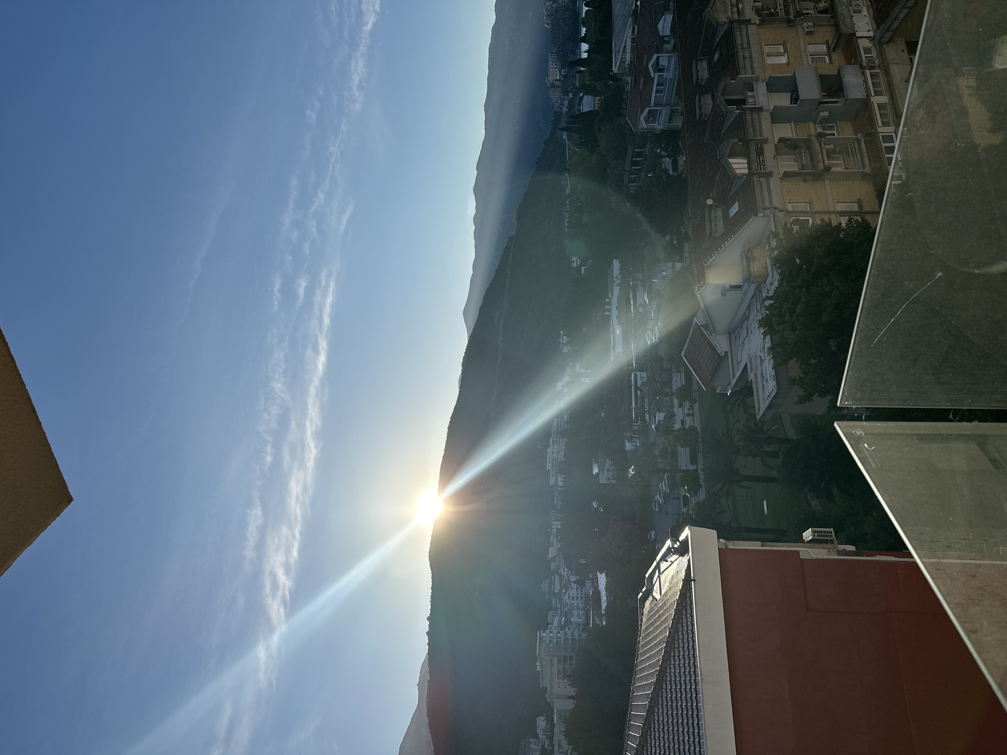 The image captures a scene in Budva, Montenegro, with the sun setting (or rising) behind a mountain range. The sky is a clear blue with wispy clouds. A lens flare from the sun is visible. The cityscape below includes buildings with orange-tiled roofs. The perspective suggests the photo was taken from a high vantage point, possibly a balcony or a high-rise building. The foreground includes part of a rooftop and glass panels.