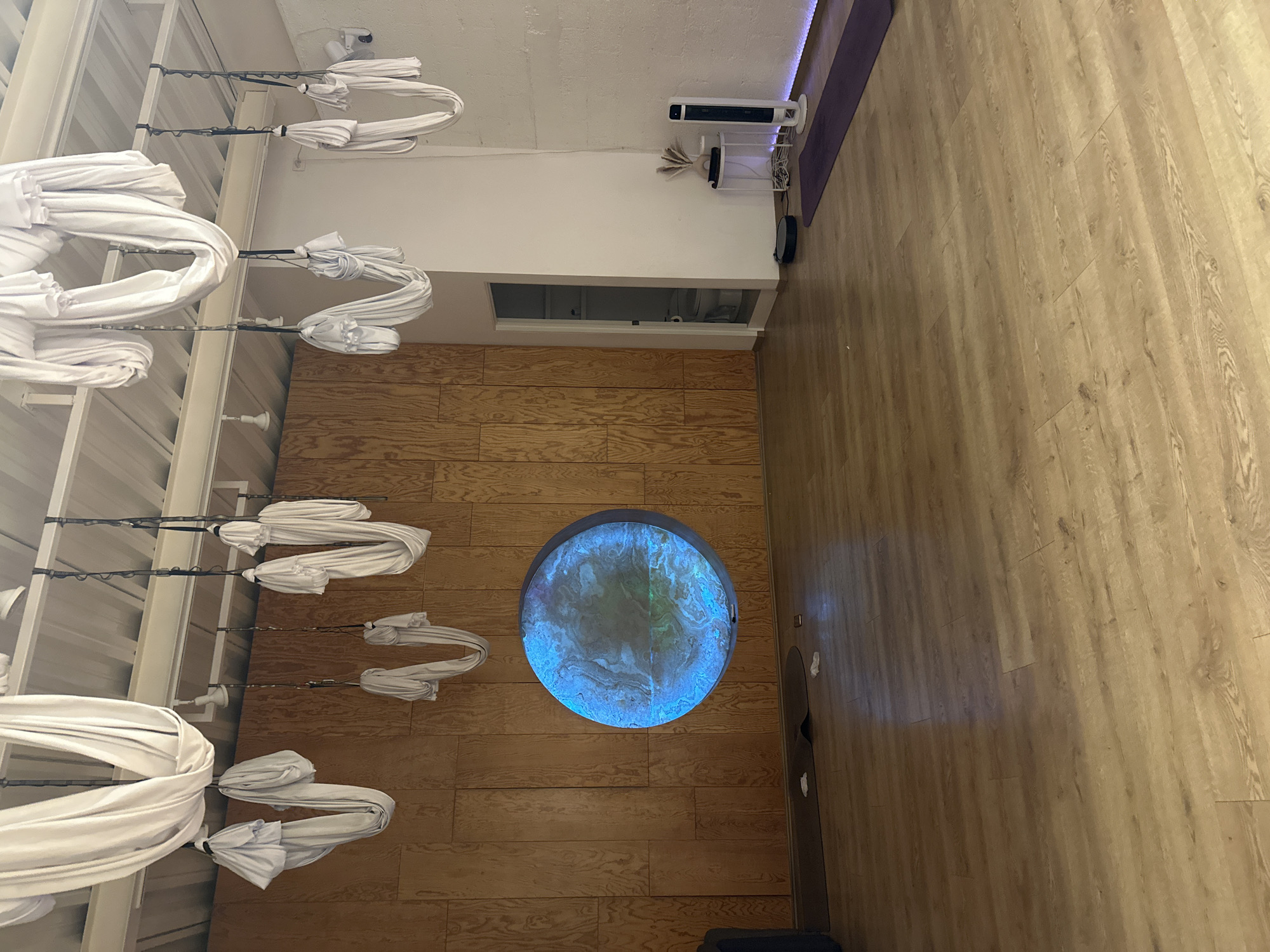 The image depicts an indoor room, likely a yoga or aerial fitness studio, viewed from a slightly elevated perspective. The room features a light-colored wooden plank floor that covers most of the visible area.
On the left side of the frame (when correctly oriented, this would be the top of the room), numerous white fabric hammocks or silks hang suspended from a metal track or ceiling structure. These silks are neatly gathered and folded, suggesting they are prepared for aerial yoga or similar activities but are not currently in use. There are approximately ten such silks visible, arranged in a row along the length of the room.
Embedded in the wooden floor, towards the center-left, is a striking circular light feature. This large disc emits a vibrant blue and greenish glow, displaying an abstract, swirling pattern that resembles a nebula or a celestial body.
Towards the right side of the room, a white low wall or partition is visible, featuring a rectangular opening or window. Above this wall, there is a plain white ceiling. Along the wall near the ceiling, several objects are placed: a white tower fan, a cylindrical white diffuser or humidifier with decorative elements, and a long, unrolled purple mat, likely a yoga mat.
No people are present in the scene, and no specific activity is taking place, but the setup clearly indicates a space designed for physical activities. The lighting in the room is bright, complemented by the internal glow of the circular floor feature. There is no visible text in the image. The environment appears clean, organized, and functional. The location context for this setting is Puebla, Mexico.