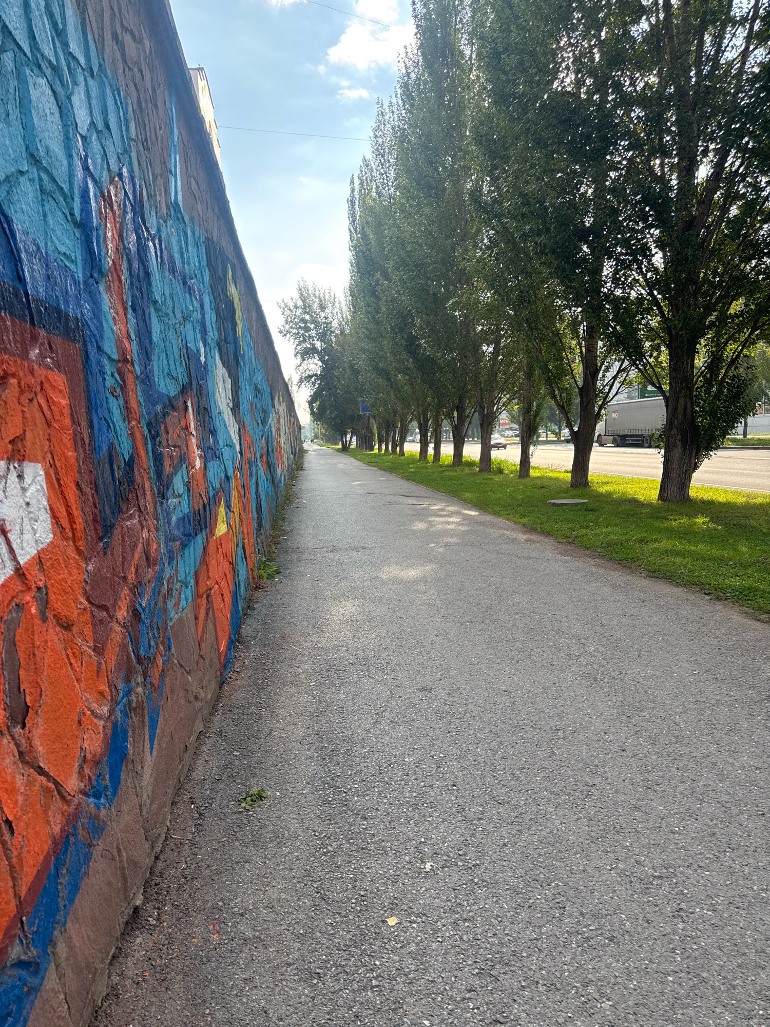 The image depicts a pedestrian walkway alongside a vibrantly graffitied stone wall, likely in Ufa, Russia. The wall, adorned with bold strokes of orange, blue, and white, dominates the left side of the frame. Beyond the wall, a line of tall, slender trees with lush green foliage forms a natural canopy, casting dappled shadows on the asphalt path. The walkway stretches into the distance, lined by a grassy verge that borders a street where a large white truck is visible. The sky above is a bright blue with scattered white clouds, suggesting fair weather. The scene appears to be a public space, possibly a park or a street adjacent to a residential or commercial area.