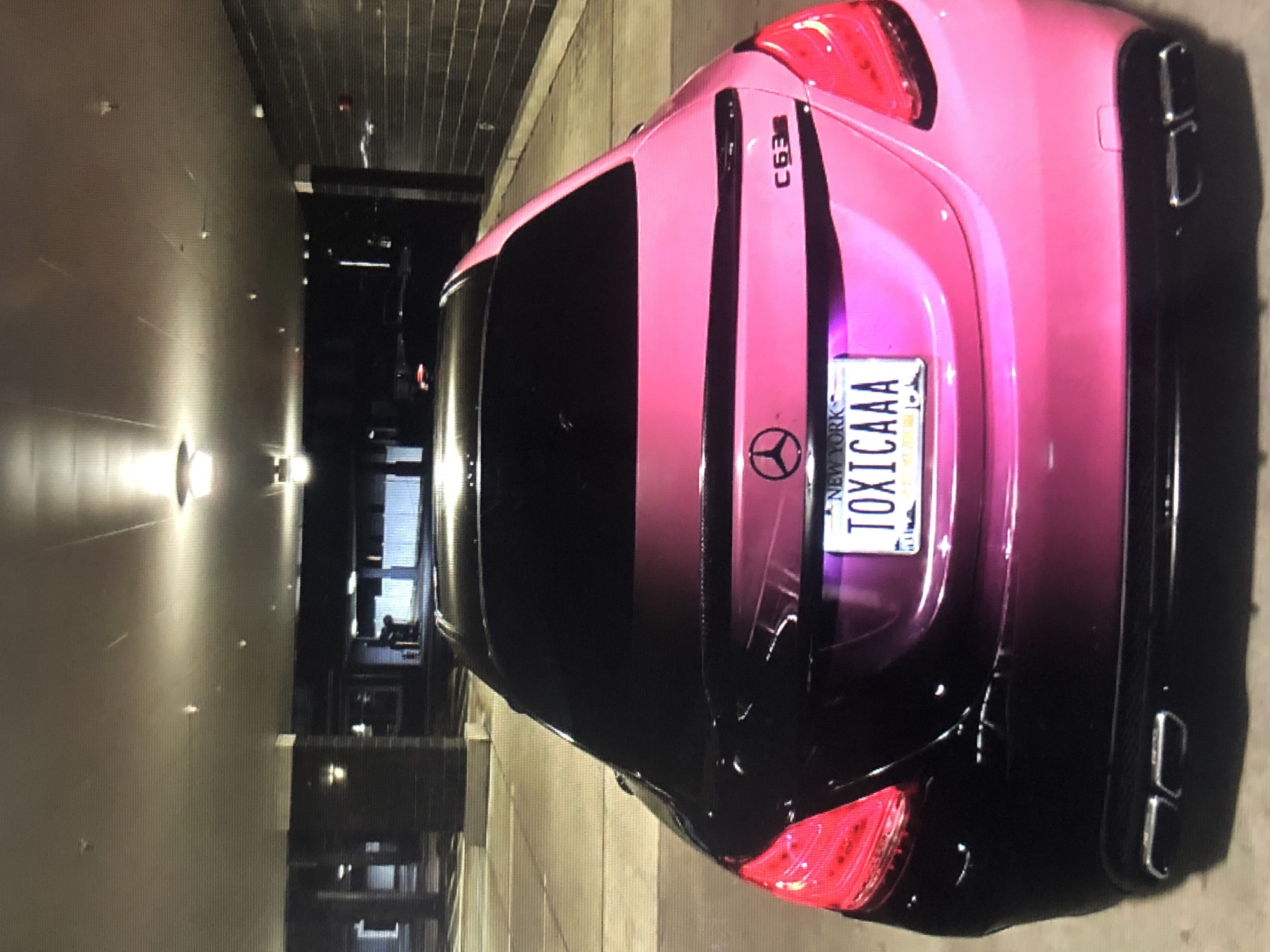 The image shows the rear of a pink and black Mercedes C63 AMG parked in an indoor parking garage. The car has a New York license plate with the word "TOXICAAA". The location is not Lhokseumawe, Indonesia, as the license plate indicates the car is registered in New York, USA. The environment is a dimly lit parking structure with fluorescent lighting visible on the ceiling. Other vehicles are faintly visible in the background.