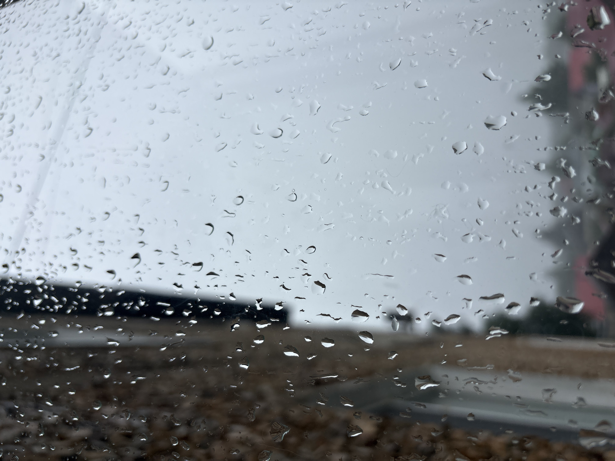 The image is a close-up view of a window covered in raindrops, with a building visible on the left side and a blurred, overcast sky and rooftops in the background. The building appears to be made of textured material, possibly brick or stone, with vertical lines suggesting window frames or architectural details. The raindrops on the glass create a distorted and abstract effect, obscuring some of the details of the exterior scene. The overall impression is one of a damp, possibly cold, day. The city of Krasnokamsk, Russia, is mentioned in the prompt, but there are no specific visual cues in the image to confirm this location. The scene is devoid of any people or discernible actions.