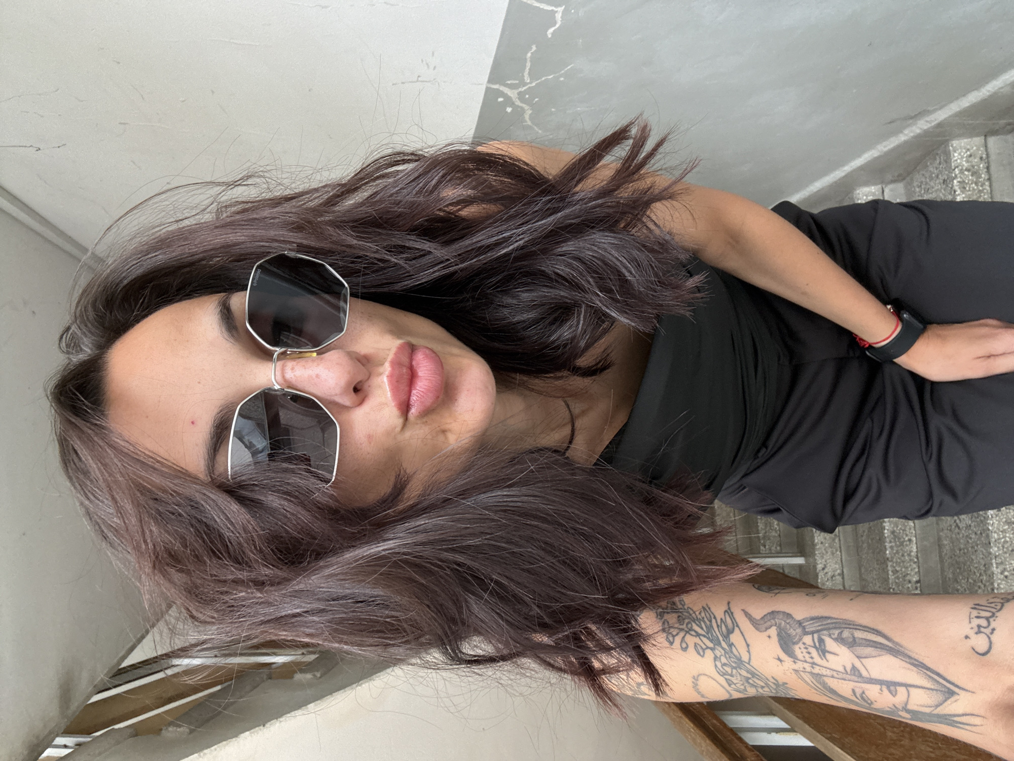 A young adult woman is pictured at an oblique angle, facing the camera, likely in an urban area within Pleven, Bulgaria. She wears dark sunglasses with silver frames, a black dress, and has tattoos on her right forearm. Her hair is dark and wavy. She has a black wristwatch with a red cord bracelet on her left wrist. The background includes a stairway with mottled steps and a cracked wall. Light appears to be entering the frame from the left side.
