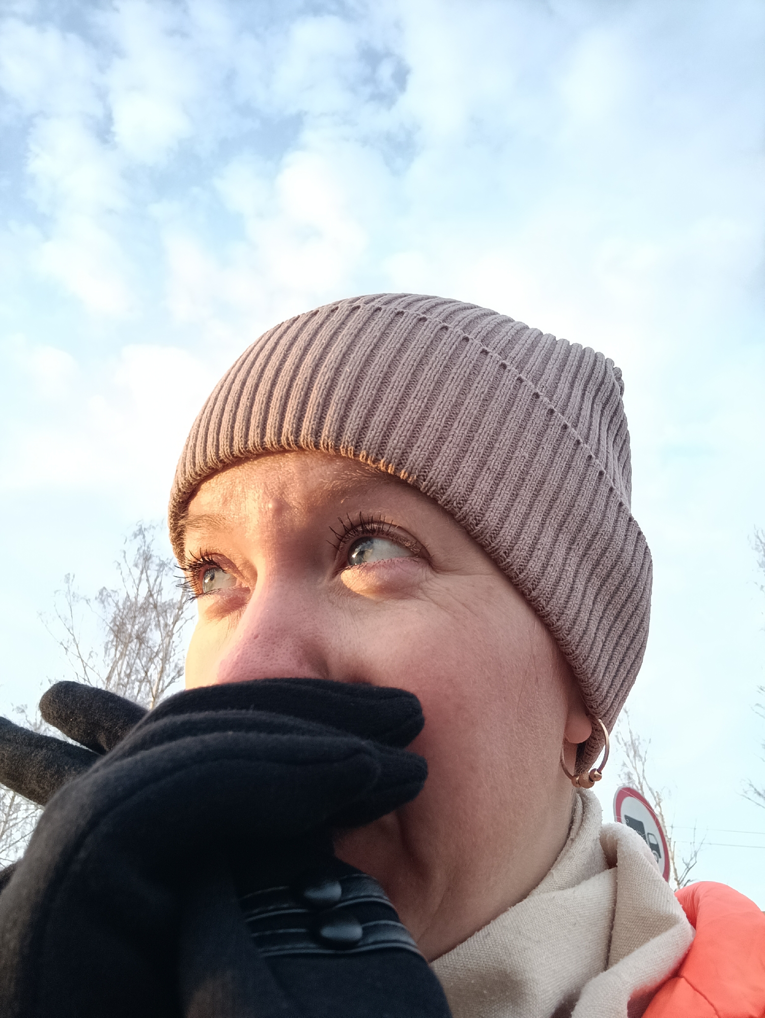 A person wearing a tan knitted hat and black gloves is looking upwards. The gloves are covering the lower part of the face. The person has light blue eyes and is wearing a hoop earring. A white scarf and orange jacket are visible. In the background are bare tree branches and a blurred road sign indicating "No Trucks". The sky is partially cloudy. Based on the information provided, this image was taken in the city of Chaikovsky, Russia.