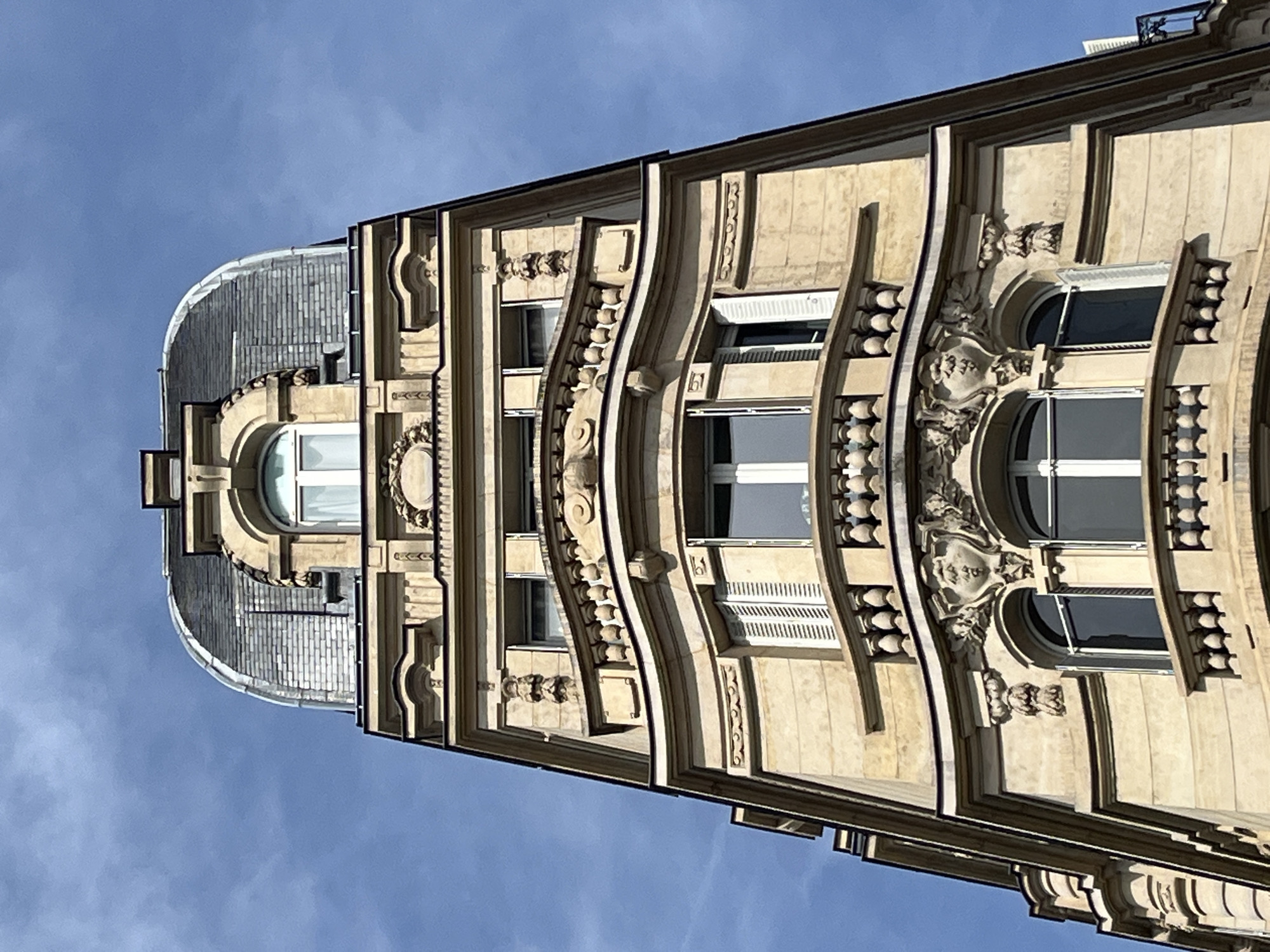 This is a low-angle, full-frame shot of the upper portion of a narrow, ornate building in Paris, France, under a clear blue sky. The building features a Mansard roof made of slate tiles, which transitions into a rounded dome at its apex. Below the roofline, the facade is made of light-colored stone and is elaborately decorated with classical architectural elements. These include arched windows, balconies with balustrades, decorative carvings above windows (including what appears to be a face), moldings, and cornices. The building's corner design is visible, with successive stories receding as they go up. There are no people or any other distinguishing features in the image.
