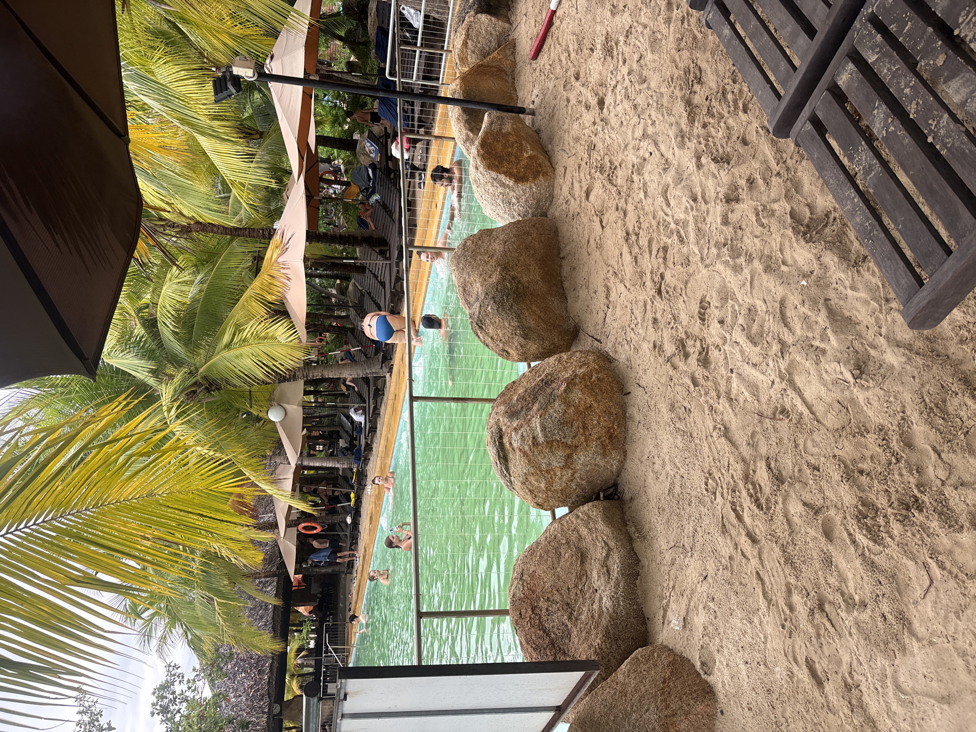 The image depicts a tropical swimming pool area with a sandy foreground. In the pool, several people are visible, some swimming and others standing. One person in the background is wearing a blue swimsuit. The pool is surrounded by large rocks and a fence. Beyond the pool, there are lounge chairs under thatched umbrellas, and numerous palm trees with vibrant green and yellow fronds. A brown umbrella shades a seating area. In the sand, there are two wooden lounge chairs in the foreground. The presence of palm trees and the beach-like sand suggests a resort or hotel in a coastal area. This scene is likely located at a resort in Nha Trang, Vietnam, known for its beaches and tropical climate.