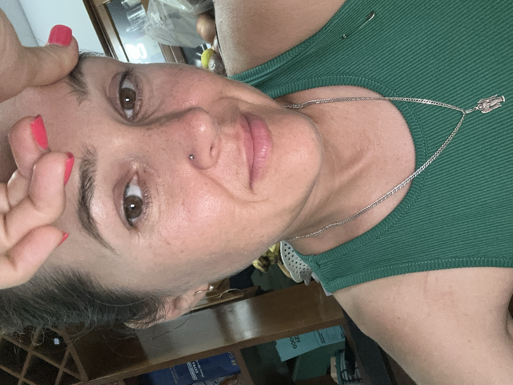A woman with a nose piercing is shown in a close-up, tilted portrait. She has brown eyes and pink nail polish. She is wearing a green ribbed tank top and a silver chain necklace with a pendant. The background includes a wooden cabinet with various objects on and inside it. The image's origin, Bac Nha Trang Ward, Vietnam, doesn't appear to be relevant to the subject.