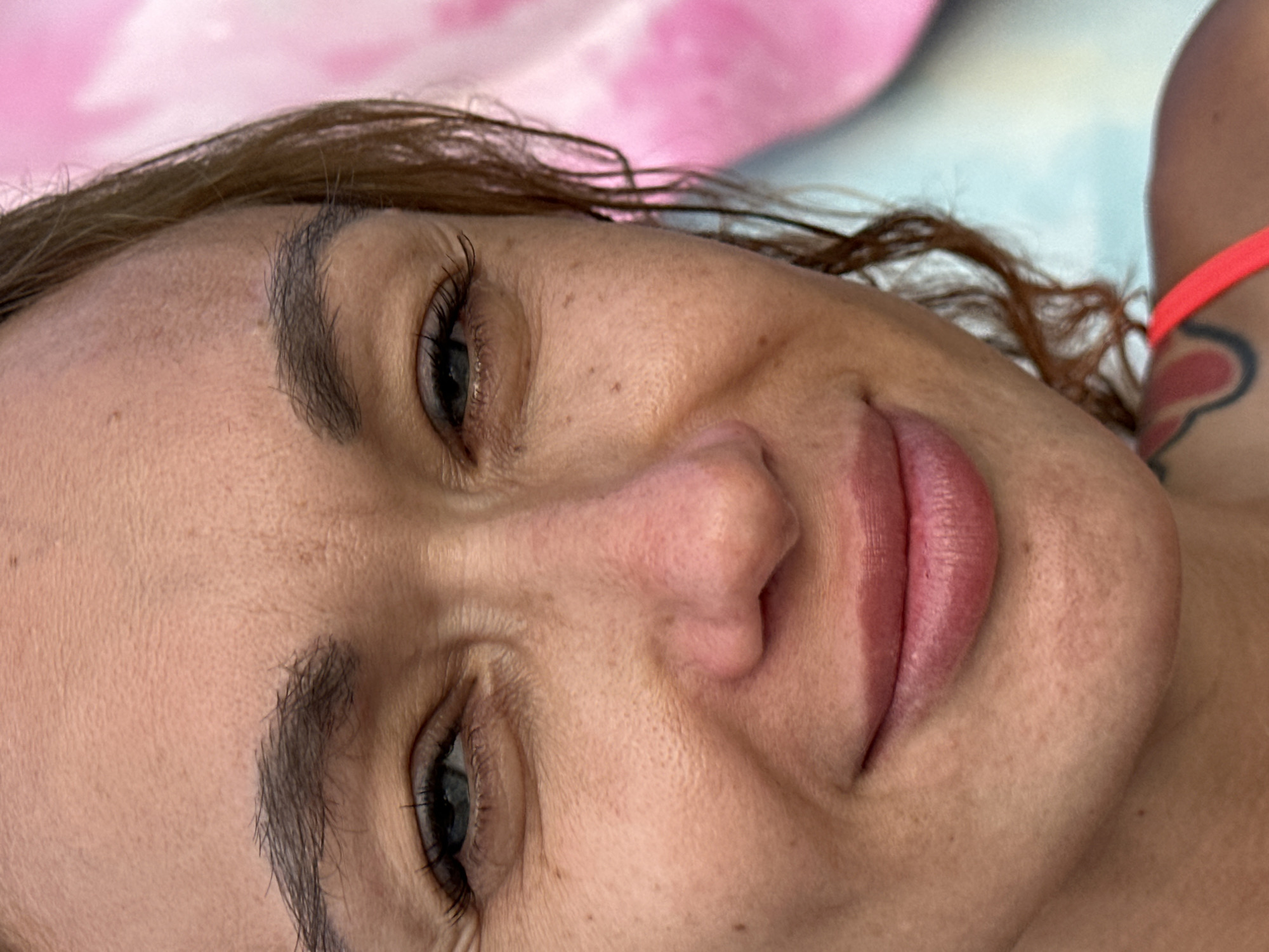 A close-up, frontal portrait shot captures the upper face and right shoulder of an adult female, located in Tien Thanh Ward, Vietnam. The subject has fair to medium skin with visible pores, freckles, and fine lines around the eyes and forehead. Her blue-grey eyes are partially closed in a subtle smile, looking directly at the viewer. Her dark eyebrows are full and well-defined. Her nose is central, with a straight bridge and a slightly rounded tip. Her full lips, colored pinkish-red, are gently upturned in a slight smile. Brown, possibly damp, hair with some curls frames the right side of her face. A bright coral-pink strap of clothing is visible on her right shoulder. A tattoo with black and red curved lines, possibly an abstract or floral design, is partially visible on her upper right shoulder/collarbone. The background is softly blurred, displaying out-of-focus areas of vibrant pink on the upper right and light blue/teal on the lower right, without discernible features.