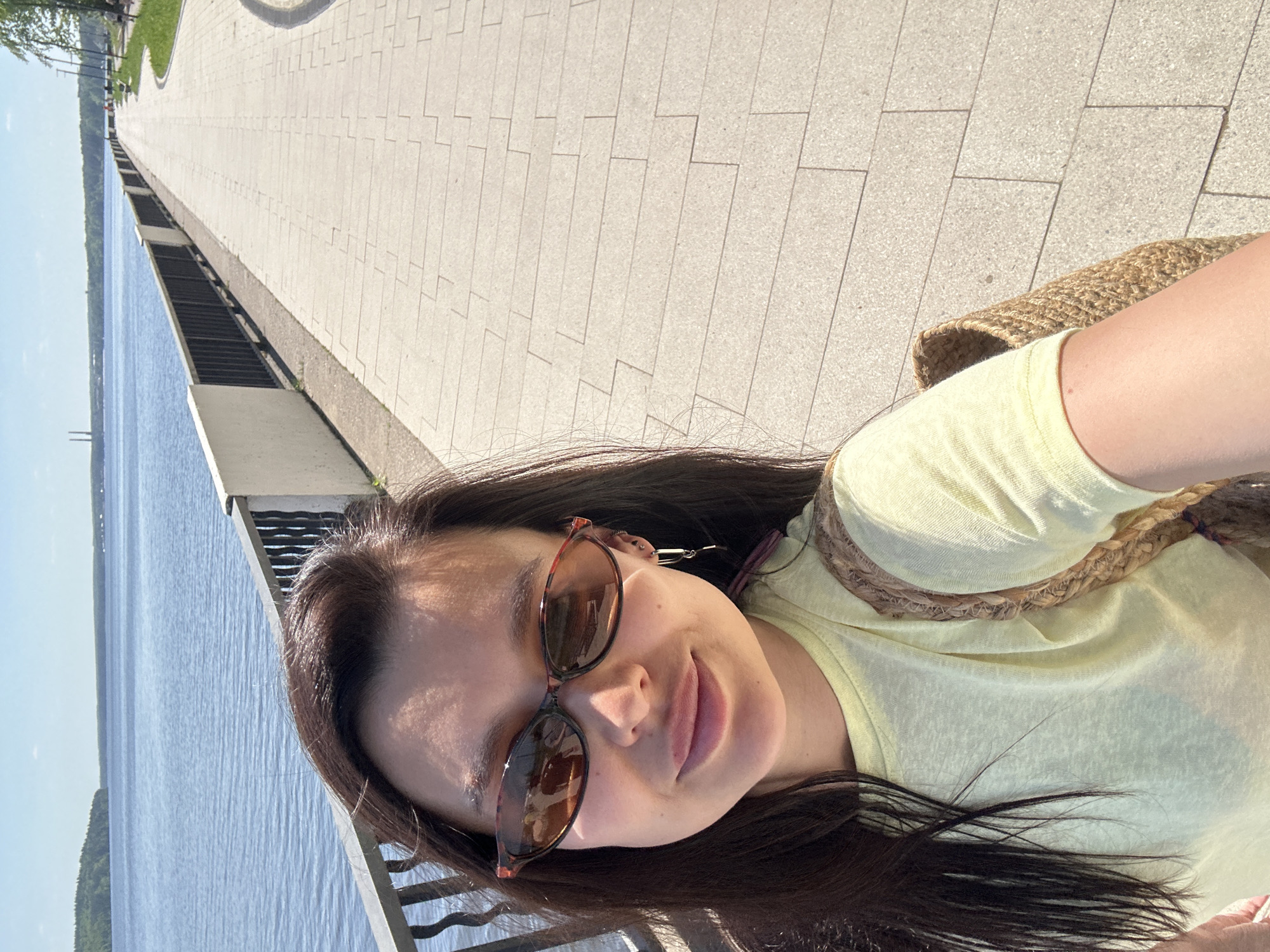 The image depicts a selfie taken by a woman with dark hair and sunglasses, wearing a yellow shirt and a straw bag. She is standing next to a body of water and a pedestrian walkway. The location is likely Chaikovsky, Russia, based on the environment. The walkway is paved with light-colored tiles, and there is a metal railing separating it from the water. The water is blue and calm, with some trees visible on the opposite shore. The weather appears sunny.
