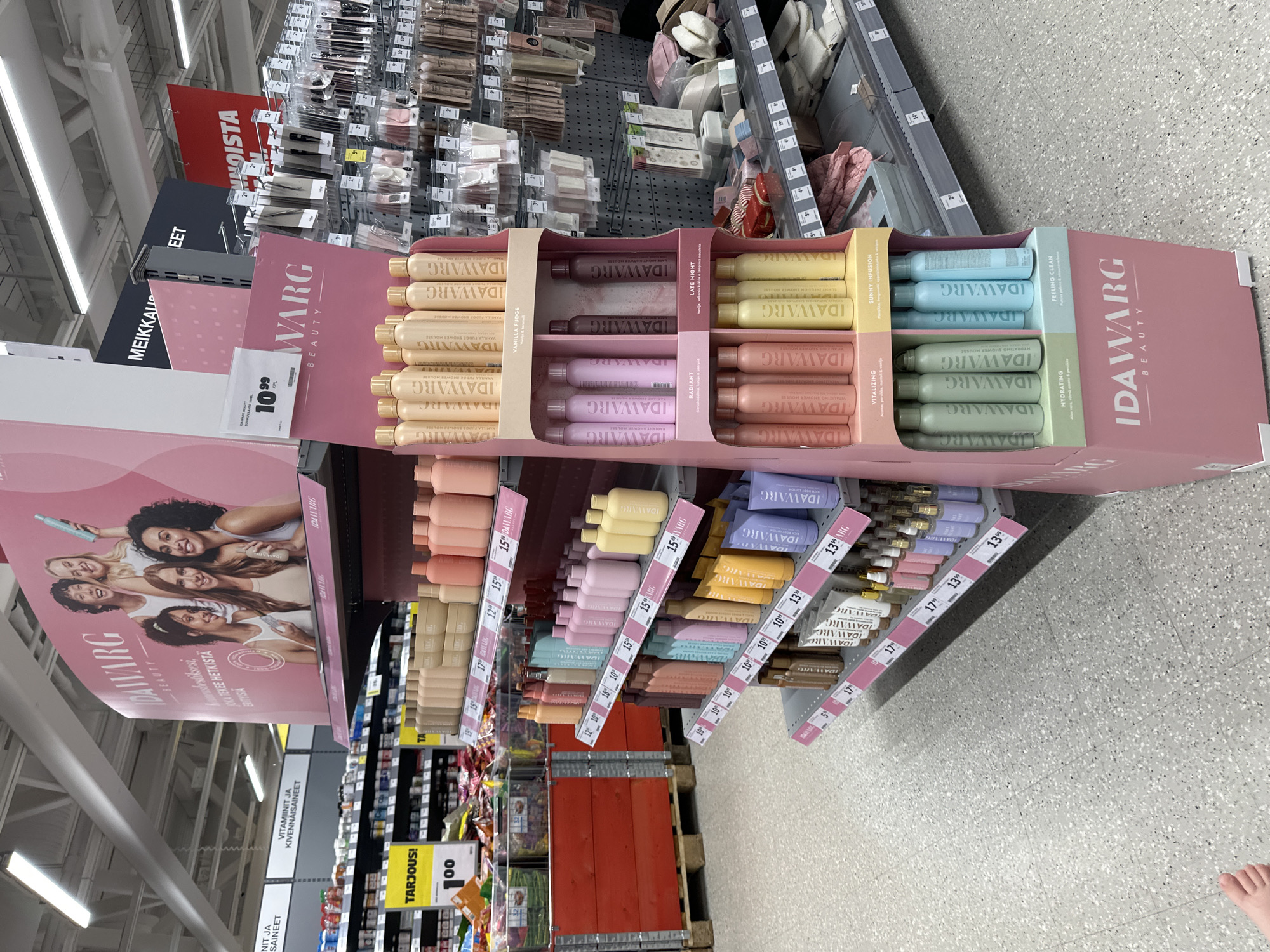 The image shows a retail store aisle with a prominent display stand featuring "IDAWARG BEAUTY" products. The stand is pink and displays various bottles and tubes of what appear to be personal care items such as shower mousse. The products have branding that includes the "IDAWARG BEAUTY" logo and flavor names like "VANILLA FUDGE", "RADIANT", "LATE NIGHT", "VITALIZING", "SUNNY INFUSION", "HYDRATING", and "FEELING CLEAN".
To the left of the display, shelves are stocked with other products, including what looks like vitamins or supplements. A yellow sign with "TARJOUS!" (Finnish for "OFFER!") and "100" is visible on one of these shelves. In the background, more shelves are filled with a variety of merchandise.
Above the display, a large pink banner with images of women and the "IDAWARG BEAUTY" logo is hung. To the right of the display, another section of the store has shelves with makeup brushes and other beauty accessories. The floor is a light-colored, speckled tile. The overall environment suggests a well-lit retail setting, possibly a supermarket or a department store. A person's foot is visible in the lower left corner of the image, indicating the presence of at least one person, though not fully captured.