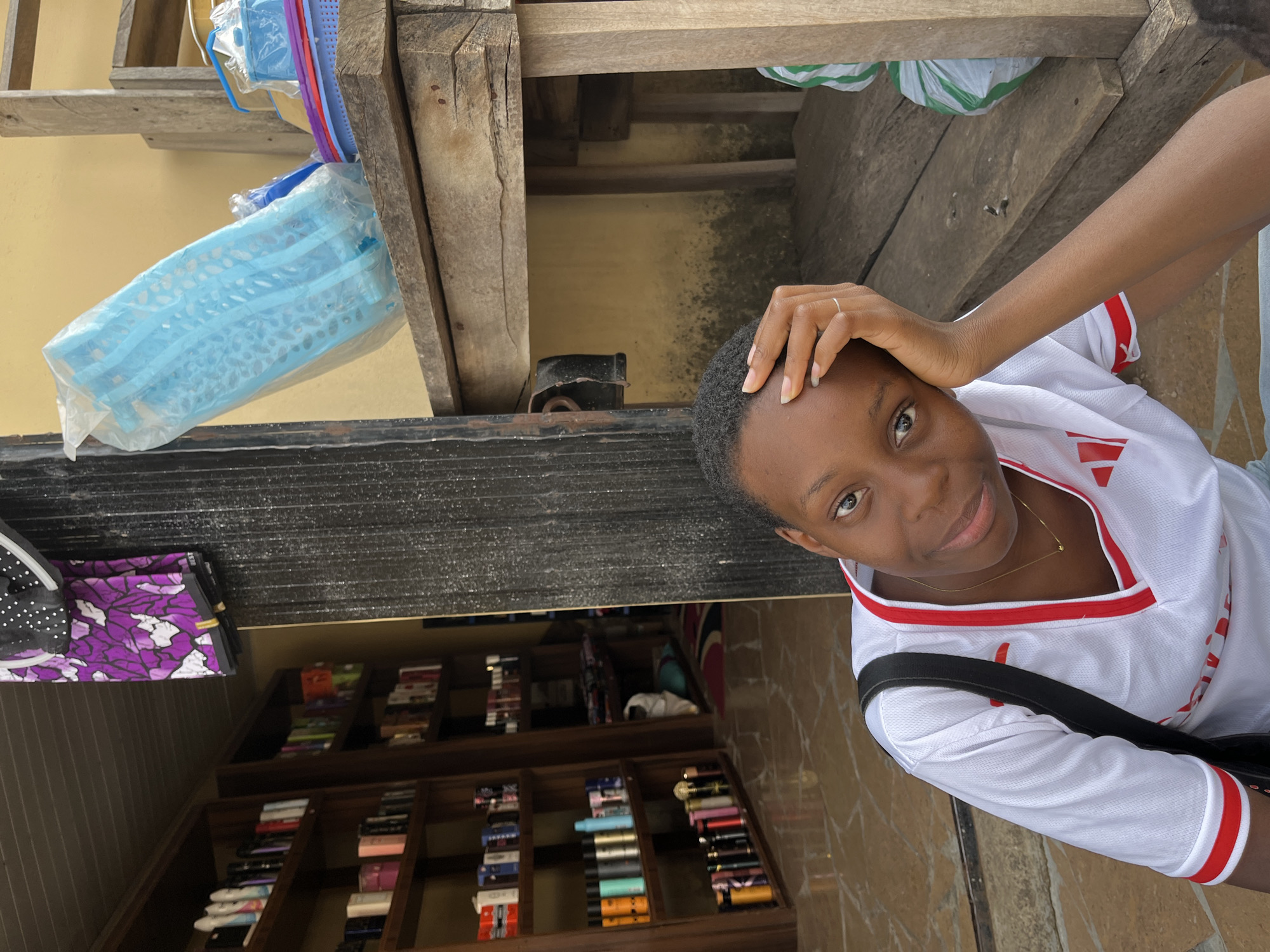 A young woman stands in front of a shop in Jos, Nigeria. She has short black hair and is wearing a white shirt with red trim. She is looking at the camera and has her hand on her forehead. A black shoulder bag is slung across her body. The shop behind her displays various items on shelves, including cosmetics and fabrics. Above, a wooden structure holds plastic storage containers, with a light yellow wall in the background.